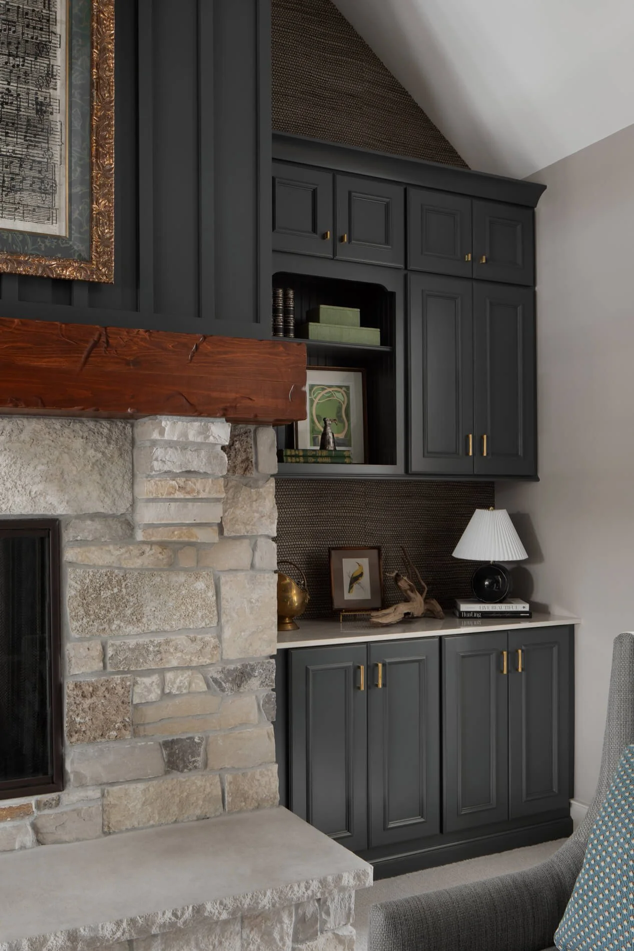 A living room corner featuring a stone fireplace with a wooden mantel, dark gray cabinetry with gold hardware, decorative objects, framed artwork, and a table lamp.