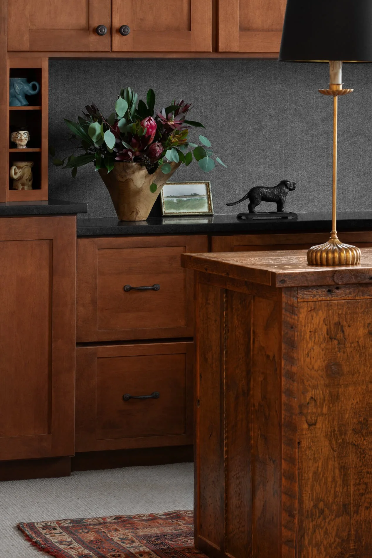 Decorative interior with wooden furniture, a large vase with fresh flowers, framed landscape painting, a dog-shaped sculpture, a tall floor lamp with a black shade, and a patterned rug.