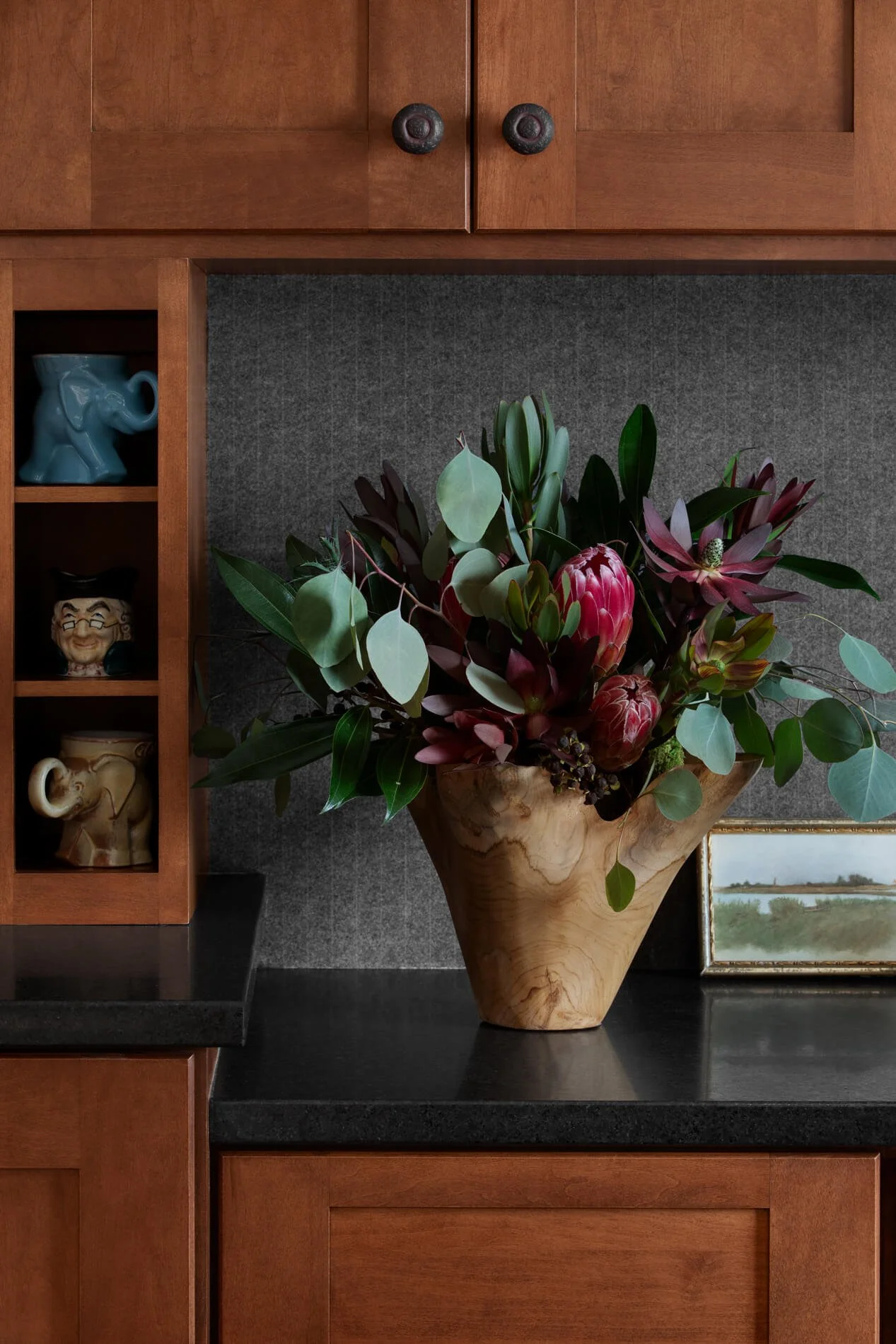 A large flower arrangement with red protea, green eucalyptus, and other foliage in a wooden vase on a black countertop.