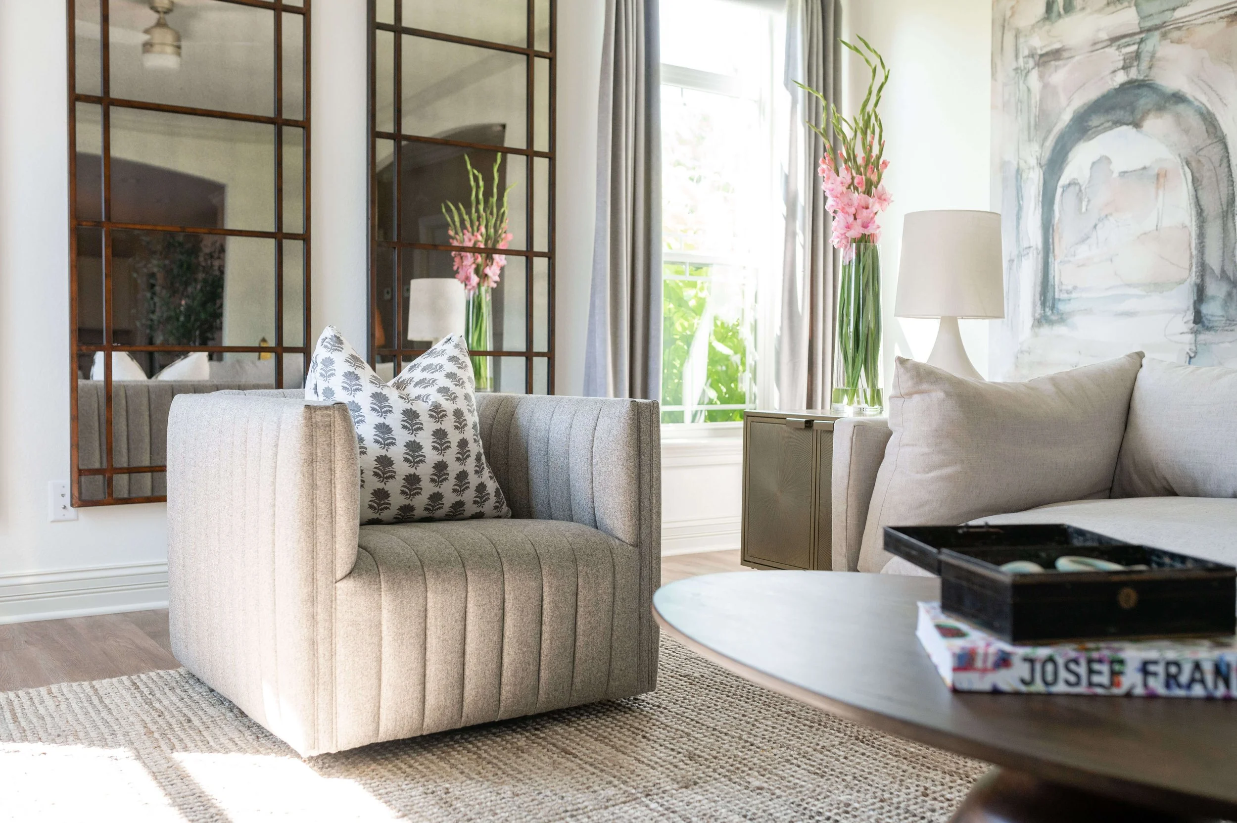 Living room with beige sofa, white pillows with black leaf pattern, glass vase with pink flowers on side table, large abstract wall art, window with curtains, mirror reflecting part of room, coffee table with books and game, lamp on side table, area rug, wooden flooring.