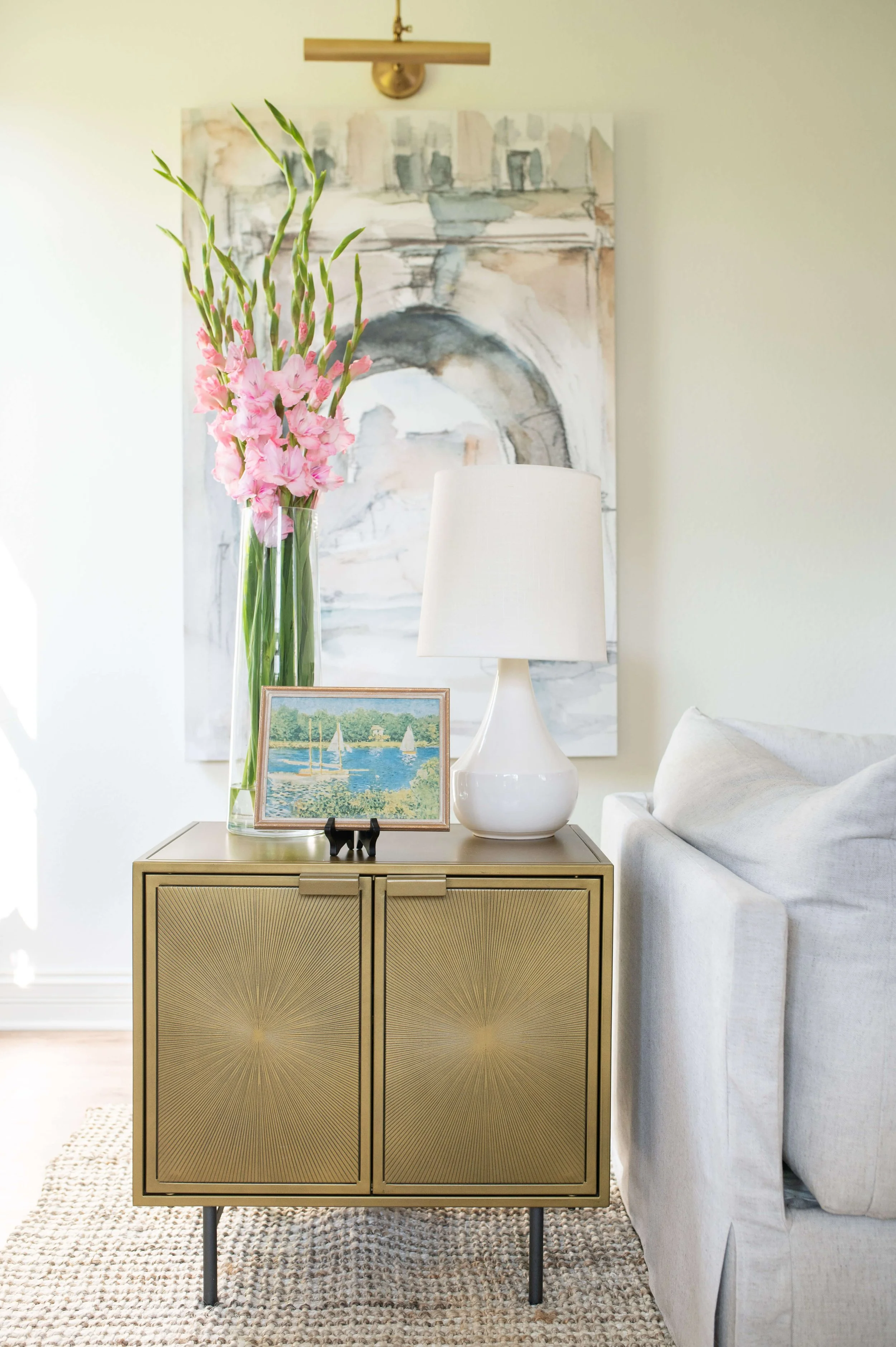 A modern living room corner featuring a gold-colored cabinet with sunburst pattern doors, a white table lamp, a framed artwork of sailboats on water, and a tall glass vase with pink gladiolus flowers. A light-colored sofa and a large abstract painting are in the background.