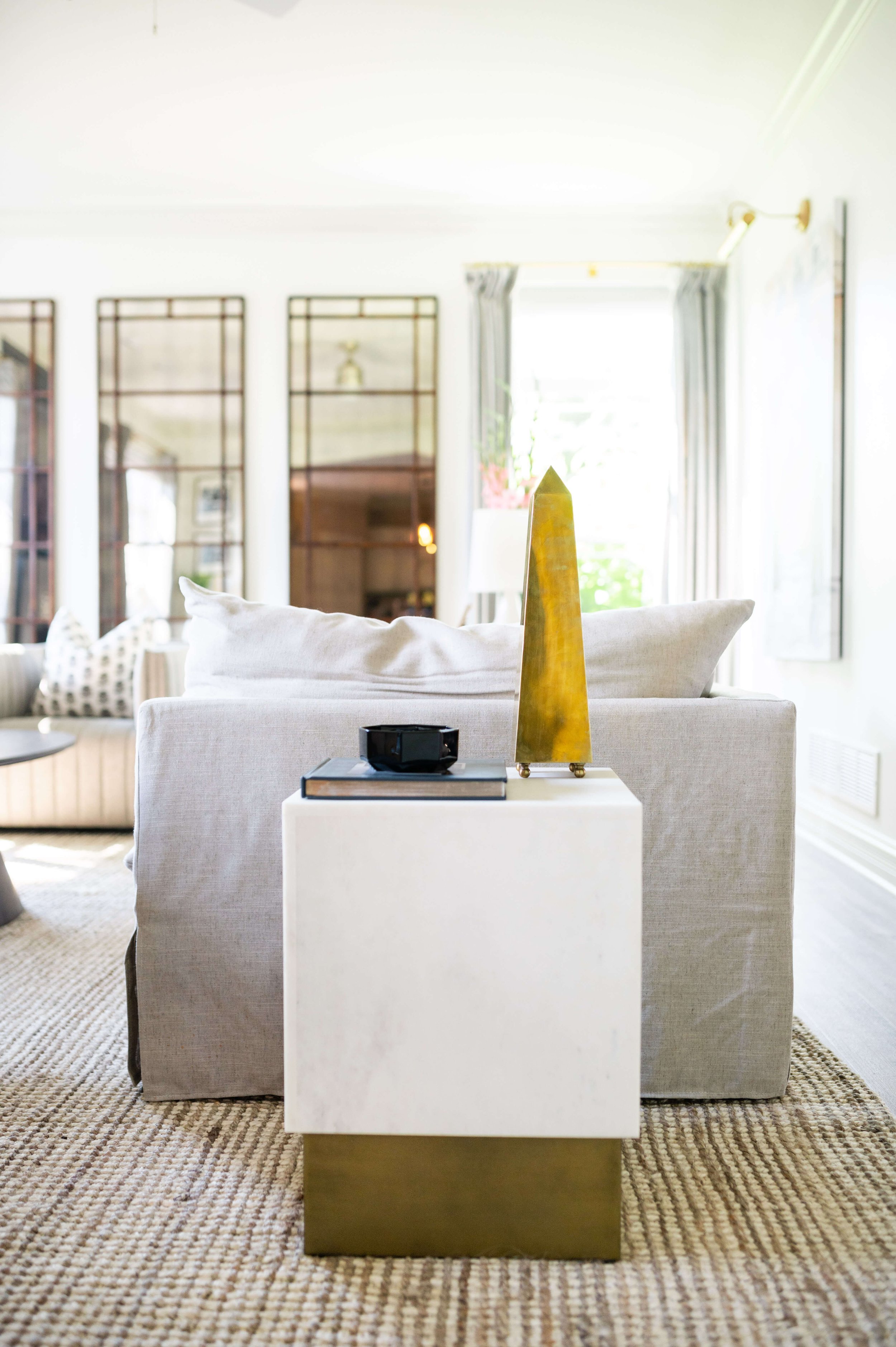 Living room with beige sofa, white cube table with a black object and a gold decorative object on top, large window with white curtains, bookshelf, and patterned rug.