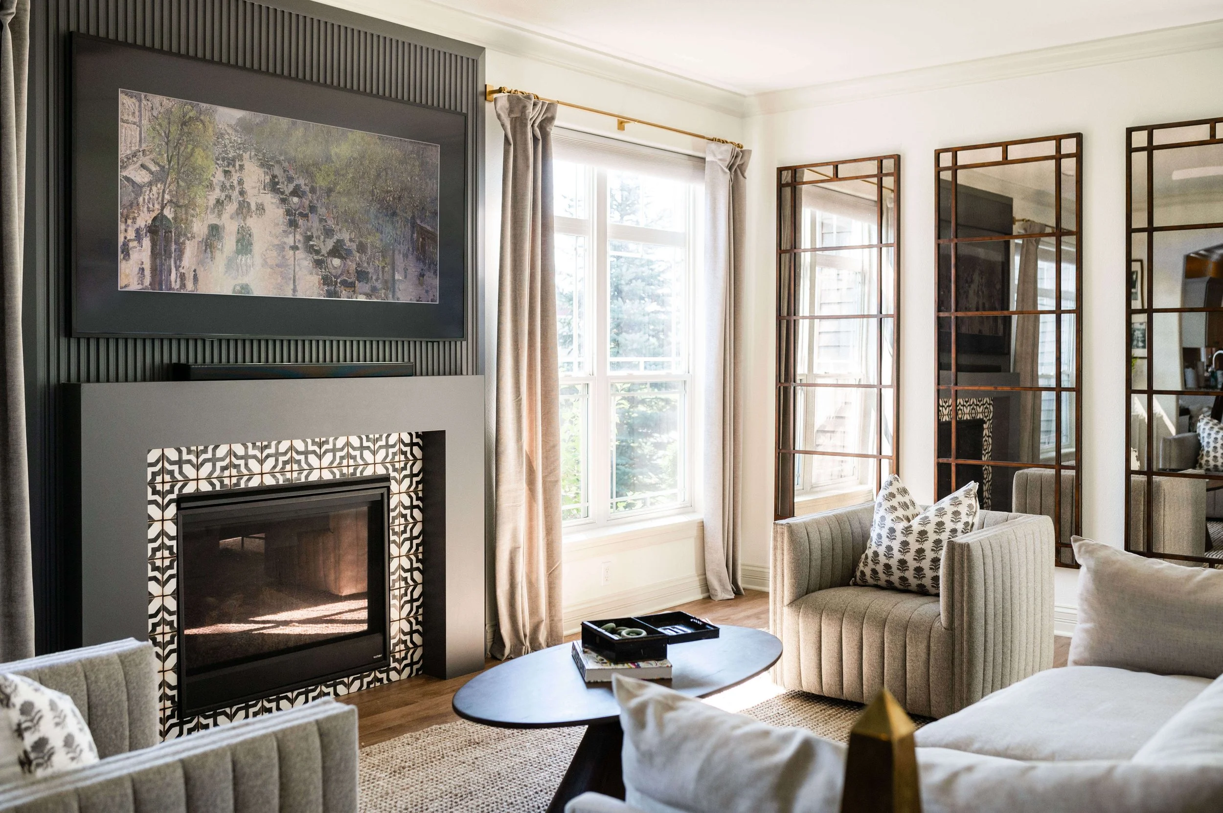 Bright living room with a patterned tile fireplace, framed artwork, large windows, mirrored walls, upholstered chairs, and a round coffee table.