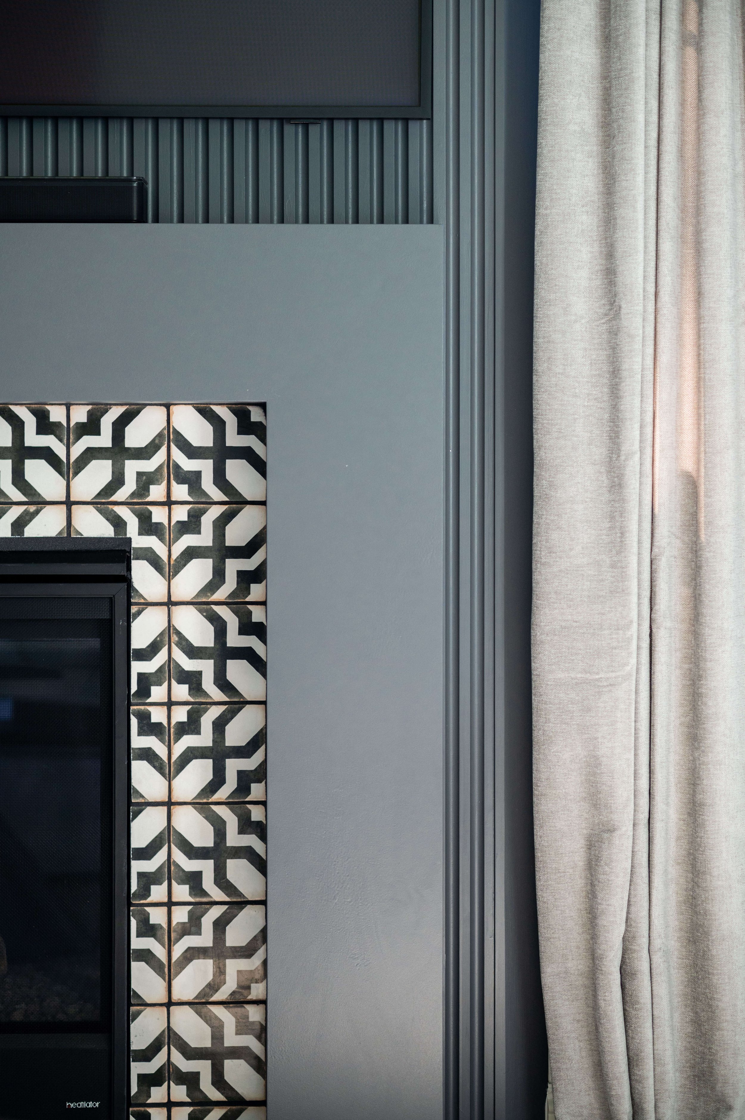 A fireplace with a black and white geometric tiled front, next to a gray wall with vertical molding, and a gray curtain on the right side.