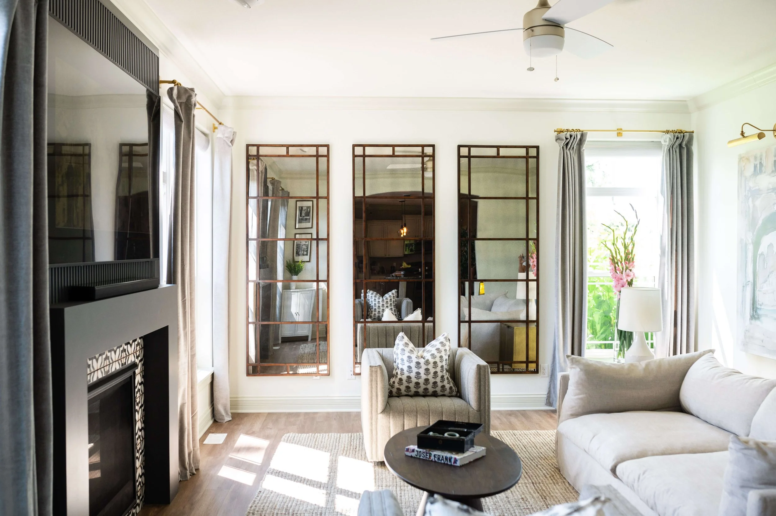 Bright living room with large windows, beige sofas, a round coffee table with books, a vase with pink flowers, a fireplace, and three large decorative mirrors on the wall. Curtains are drawn to the side, and sunlight streams in.