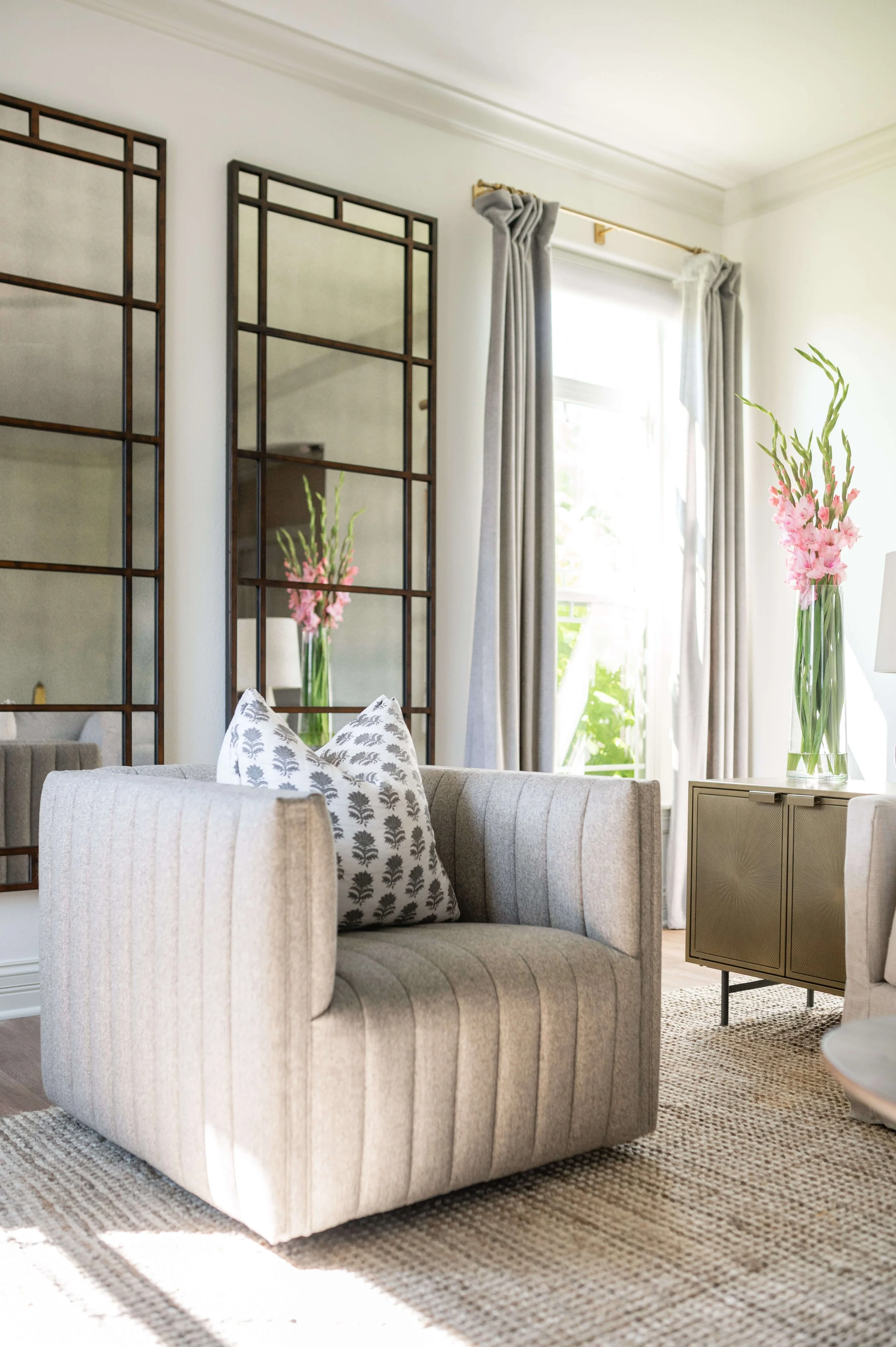 A living room with a beige upholstered armchair, a patterned throw pillow, tall glass vases with pink gladiolus flowers, and a beige cabinet, with large windows and gray curtains letting in natural light.