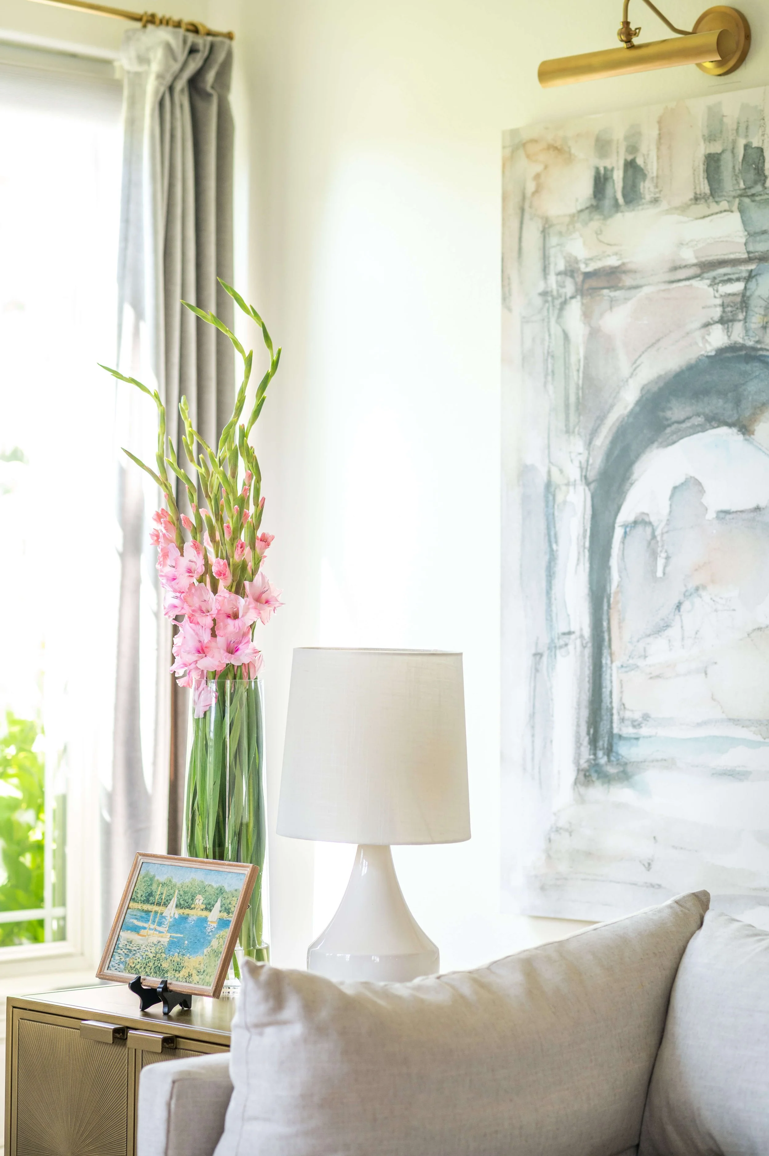 Living room with a large window, beige curtains, a tall glass vase with pink gladiolus flowers, a framed landscape painting, a white table lamp, a beige sofa with cushions, and abstract art on the wall.