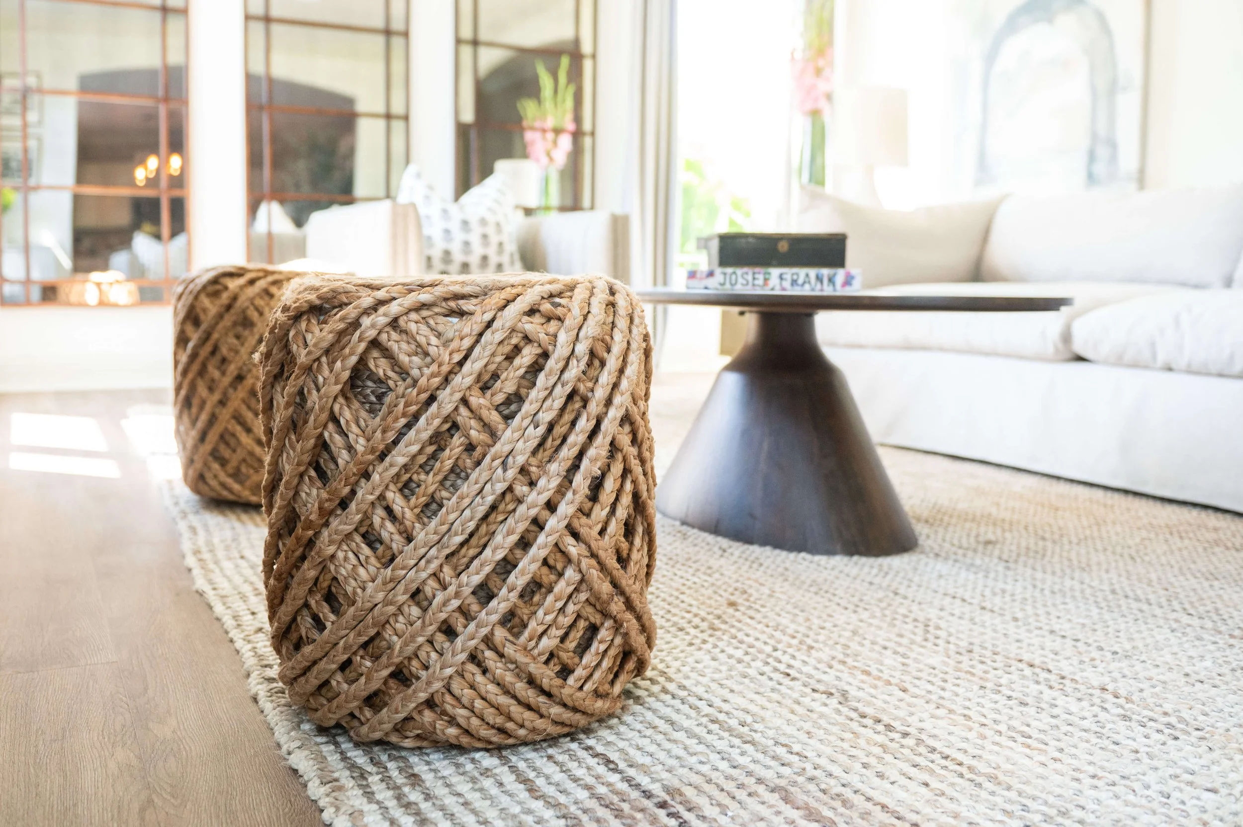 Close-up of a woven wicker basket on a beige and brown striped rug in a sunlit living room with white furniture and large windows.