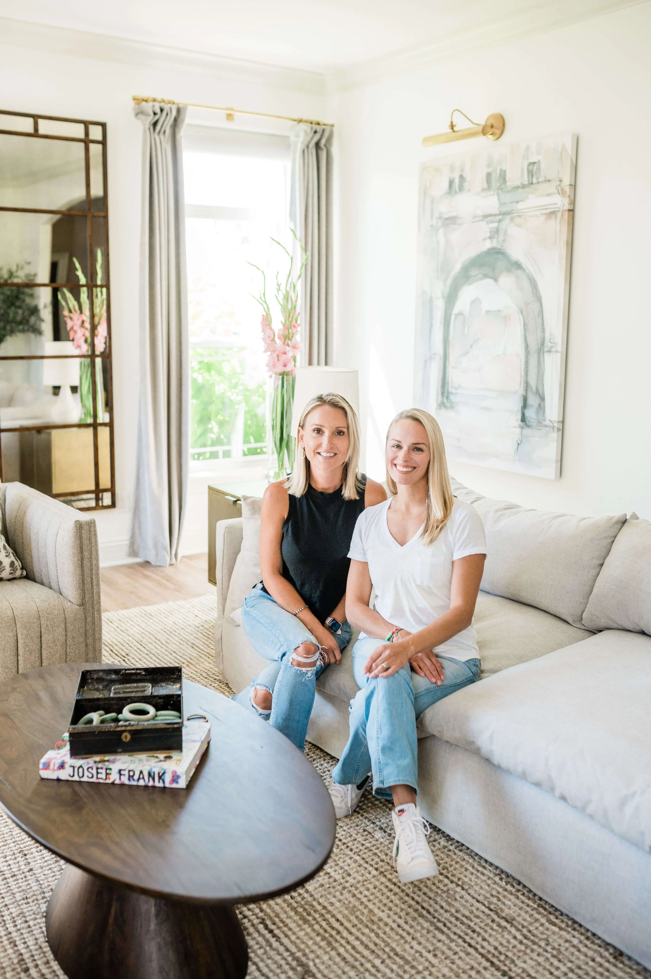 Two blonde women sit on a light-colored sofa in a bright, modern living room, smiling at the camera.
