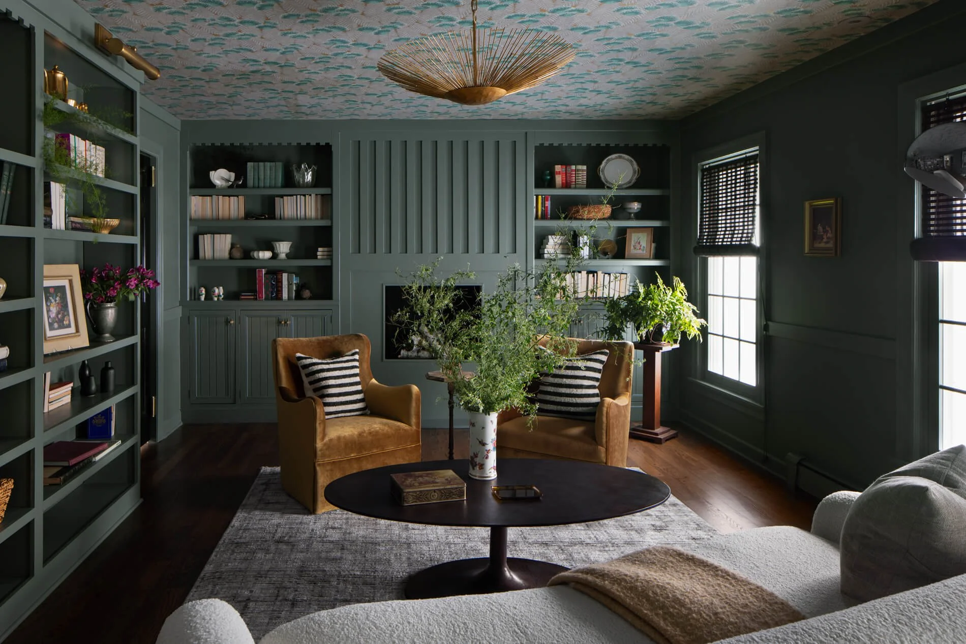 Living room with dark green walls, built-in bookshelves, two windows with dark blinds, and a ceiling with colorful patterned wallpaper. There are two mustard-colored armchairs with striped pillows around a dark round coffee table, which has a book and a mobile phone on it. A large vase with green foliage is in the center of the room. There is a light-colored sofa in the foreground and a gray area rug on the dark hardwood floor.