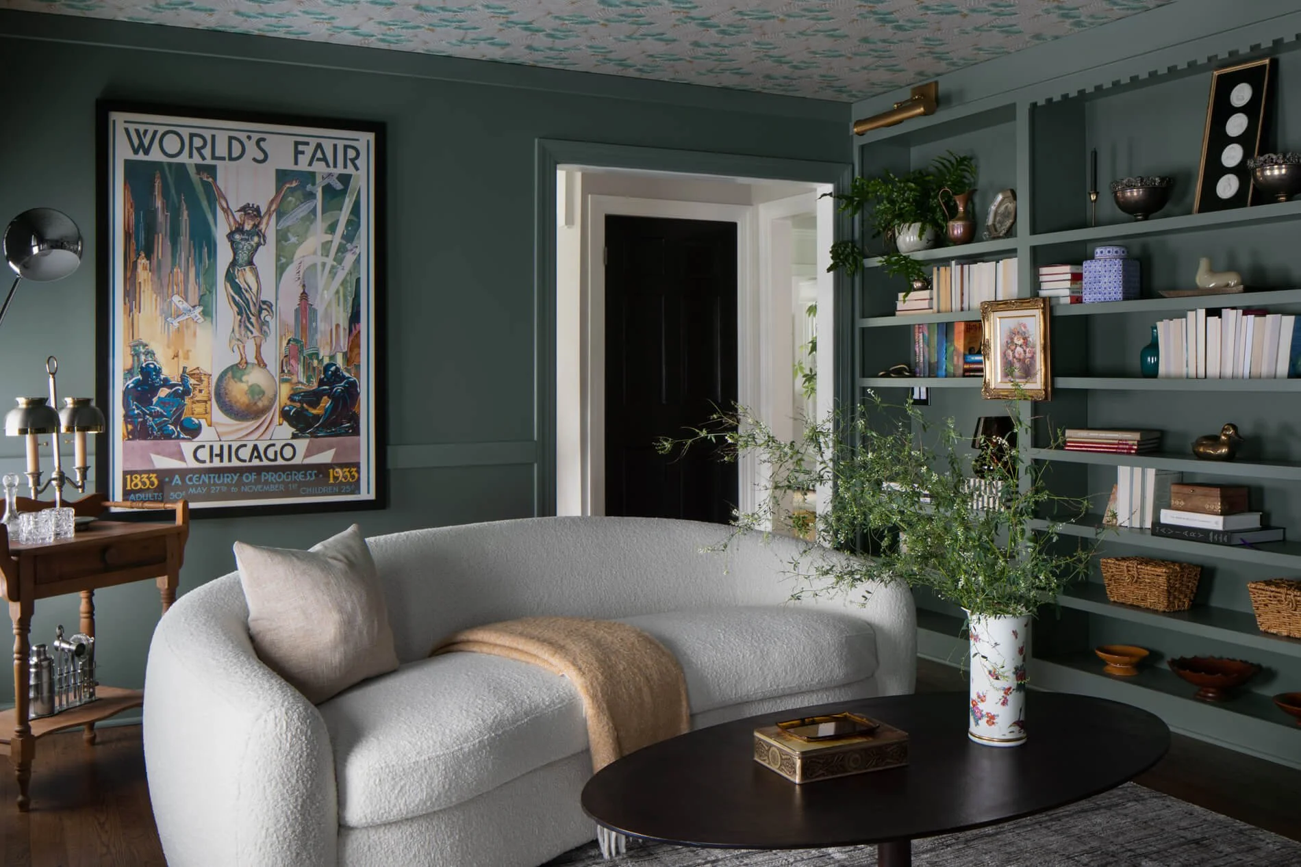 Elegant dark green living room with a white curved sofa, built-in bookshelves, floral arrangement, coffee table, and vintage 1933 Chicago World's Fair poster