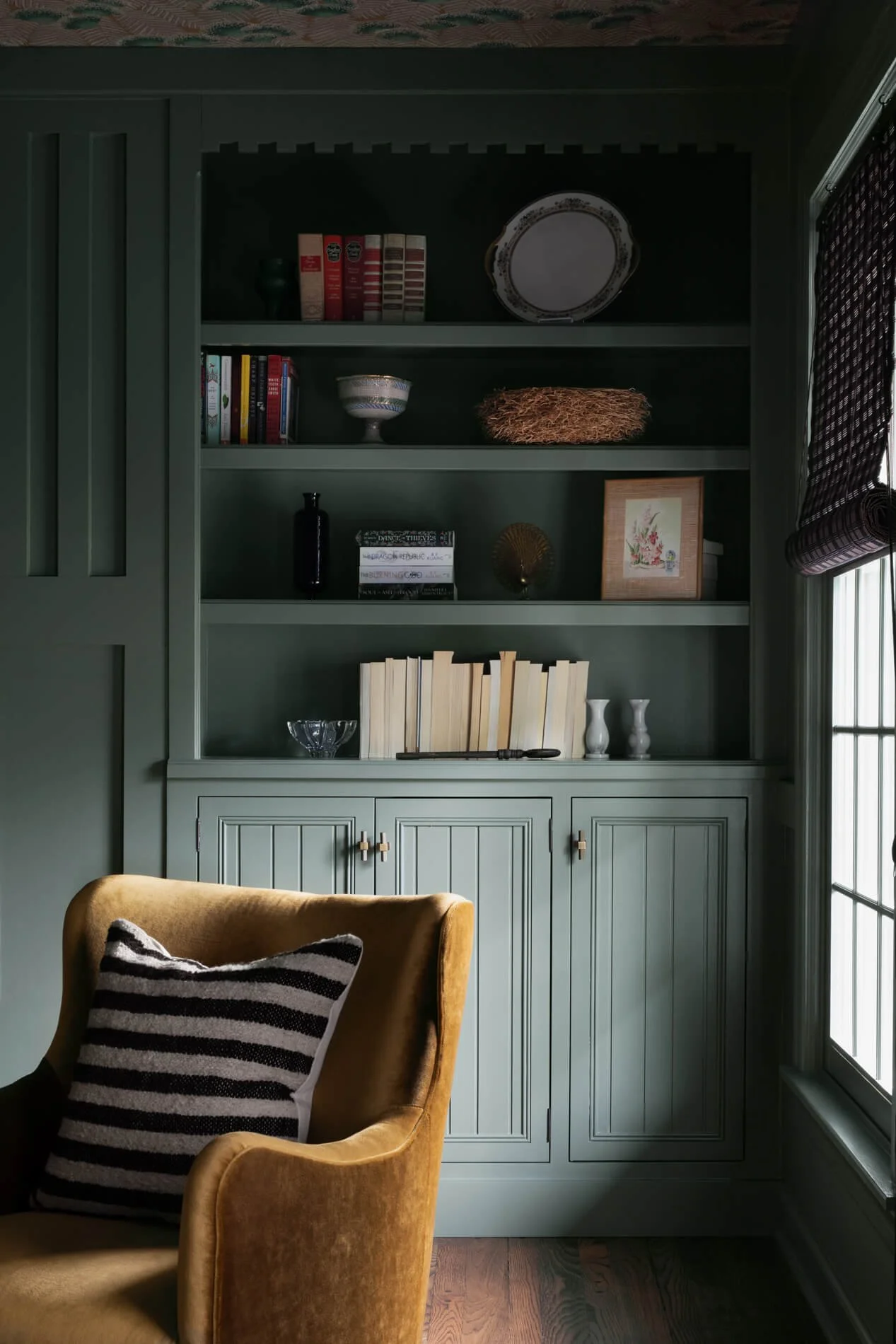 A green built-in bookshelf with three shelves containing books, decorative plates, vases, and ornaments. Part of a window with a dark patterned Roman shade is visible on the right. In front of the shelf is a tan upholstered armchair with a black and white striped pillow on it.