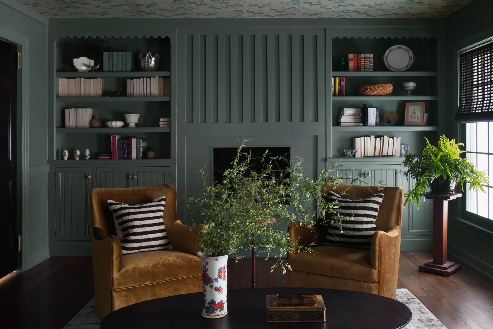 Living room with green built-in shelves, a brick fireplace, two mustard-colored armchairs with striped pillows, a black coffee table with a floral vase, and plants near a window with black blinds.