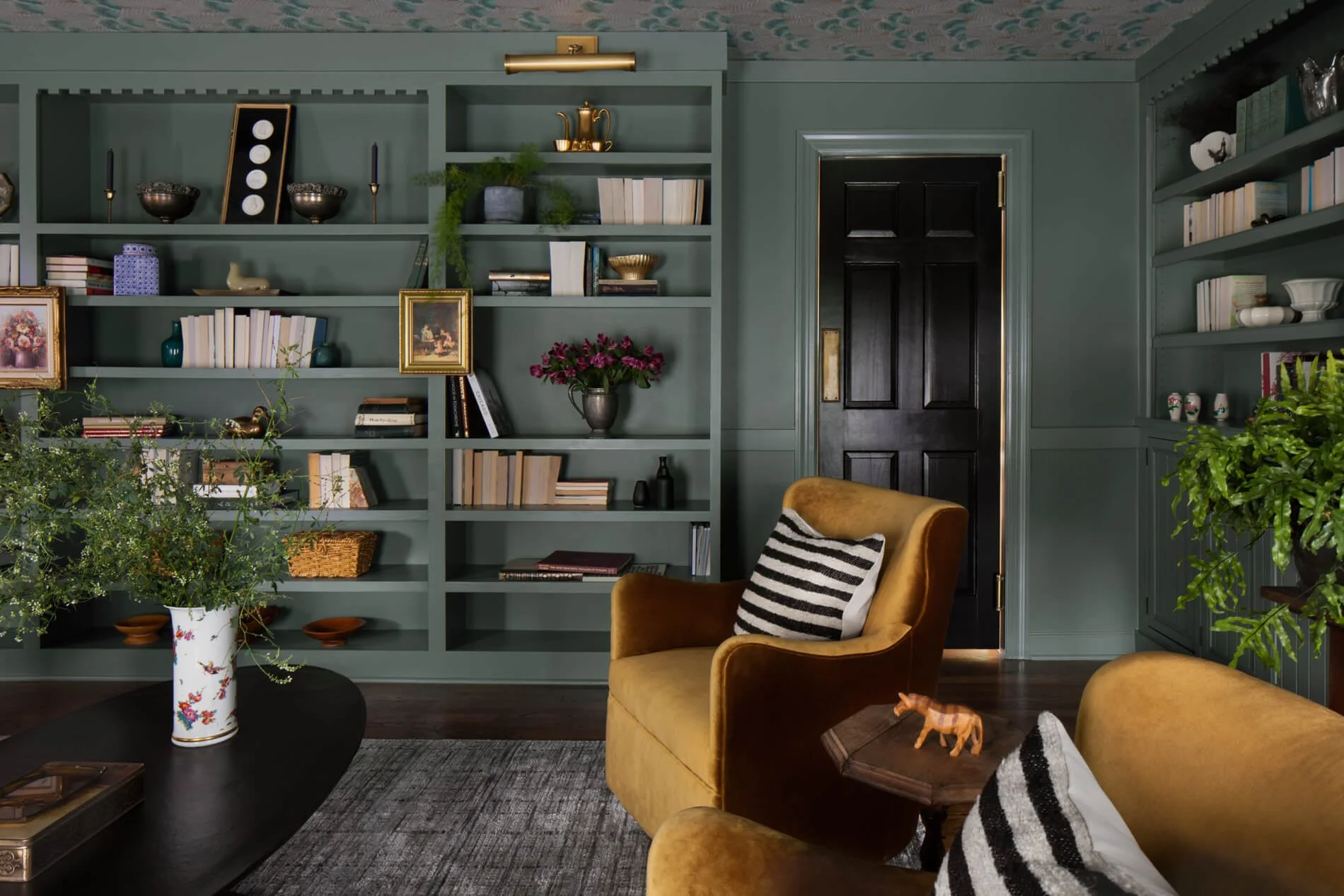 A cozy living room with a black door, an olive-green bookshelf filled with books, decorative bowls, and framed artwork. There is a mustard yellow armchair in the foreground with a pillow, and a large floral vase with greenery on a black coffee table.
