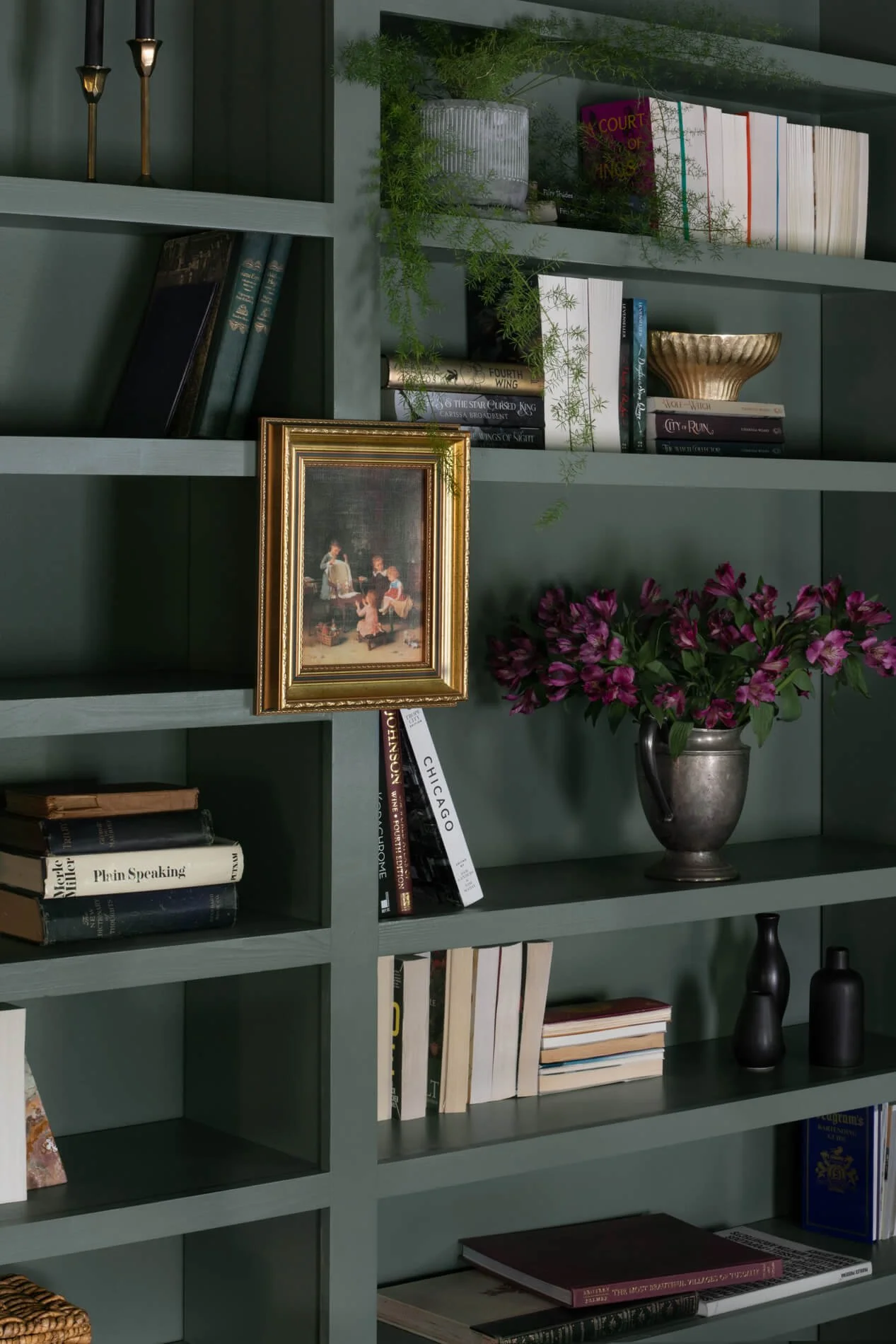 Green bookshelf with various books, a framed painting, green plants, and decorative vases and candles.