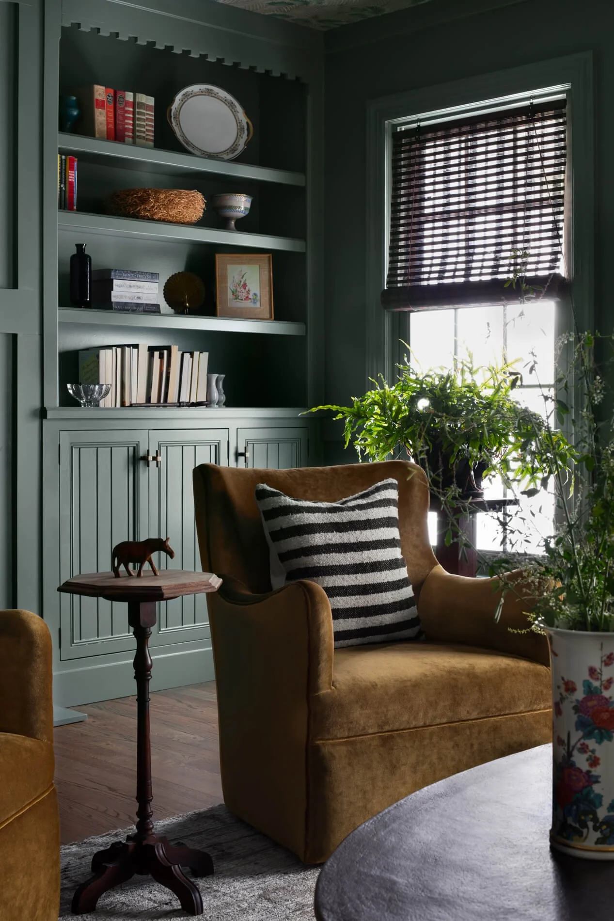 A cozy living room with a yellow armchair, striped pillow, houseplant, wooden side table with a small horse figurine, a dark round table, a green bookshelf with decorative items and books, a window with dark striped blinds, and wooden flooring.