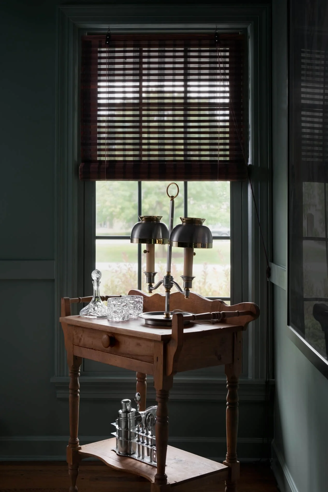 A kitchen scene featuring a window with wooden blinds, a wooden table underneath, and a brass chandelier-style light fixture on the table. The table has glassware and a metal drink caddy.