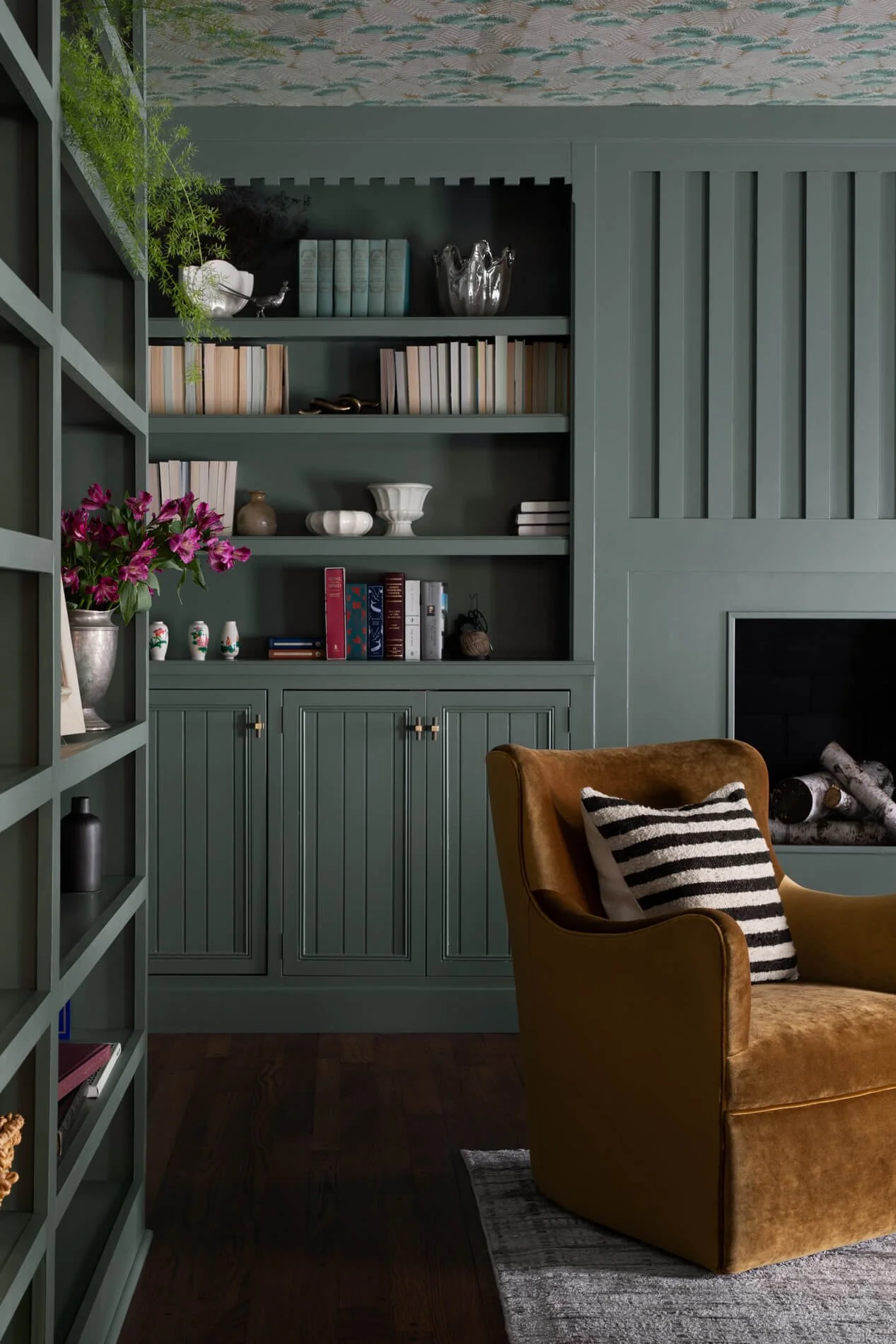 A cozy living room with a tan armchair with a black and white striped pillow, a dark green bookshelf filled with books and decorative objects, a fireplace with logs, and a patterned ceiling.
