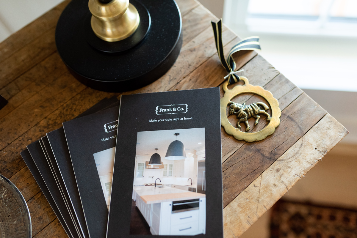 Brochures for Frank & Co. featuring home interior design, a brass and black decorative lion ornament with a ribbon, brass candlestick holder, and a wooden table.