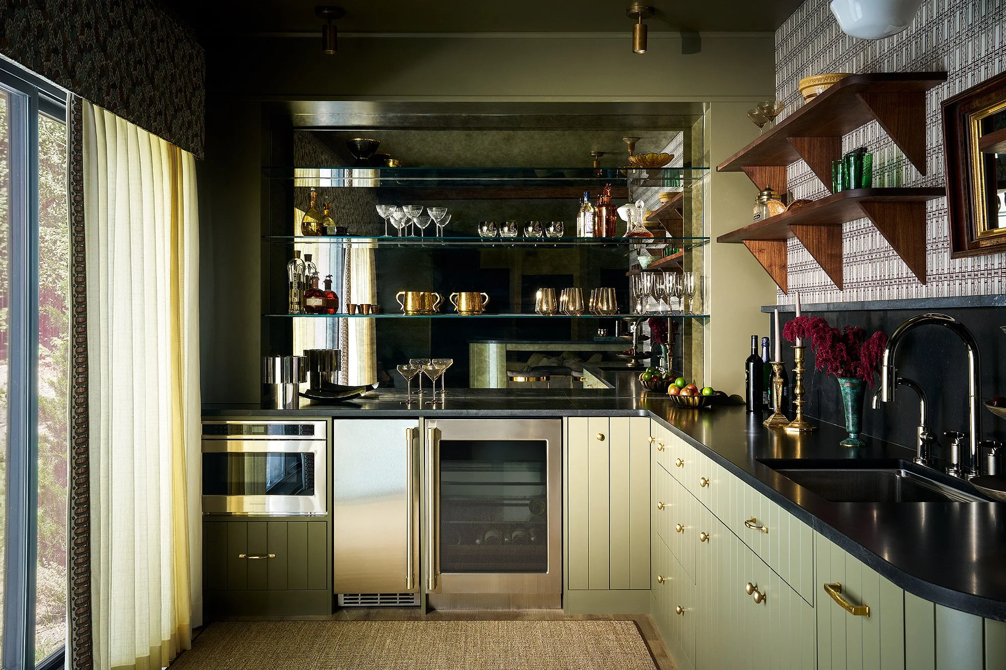 Modern kitchen featuring white cabinets with gold knobs, black countertop, built-in appliances, glass shelves with glassware and decorative items, window with light curtains, and various kitchen accessories on the counter.