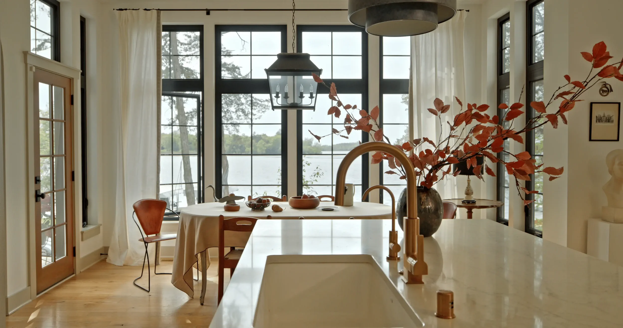 Bright lakeside dining room and kitchen with large black-framed windows, white curtains, farmhouse table, pendant lights, and autumn foliage in a vase