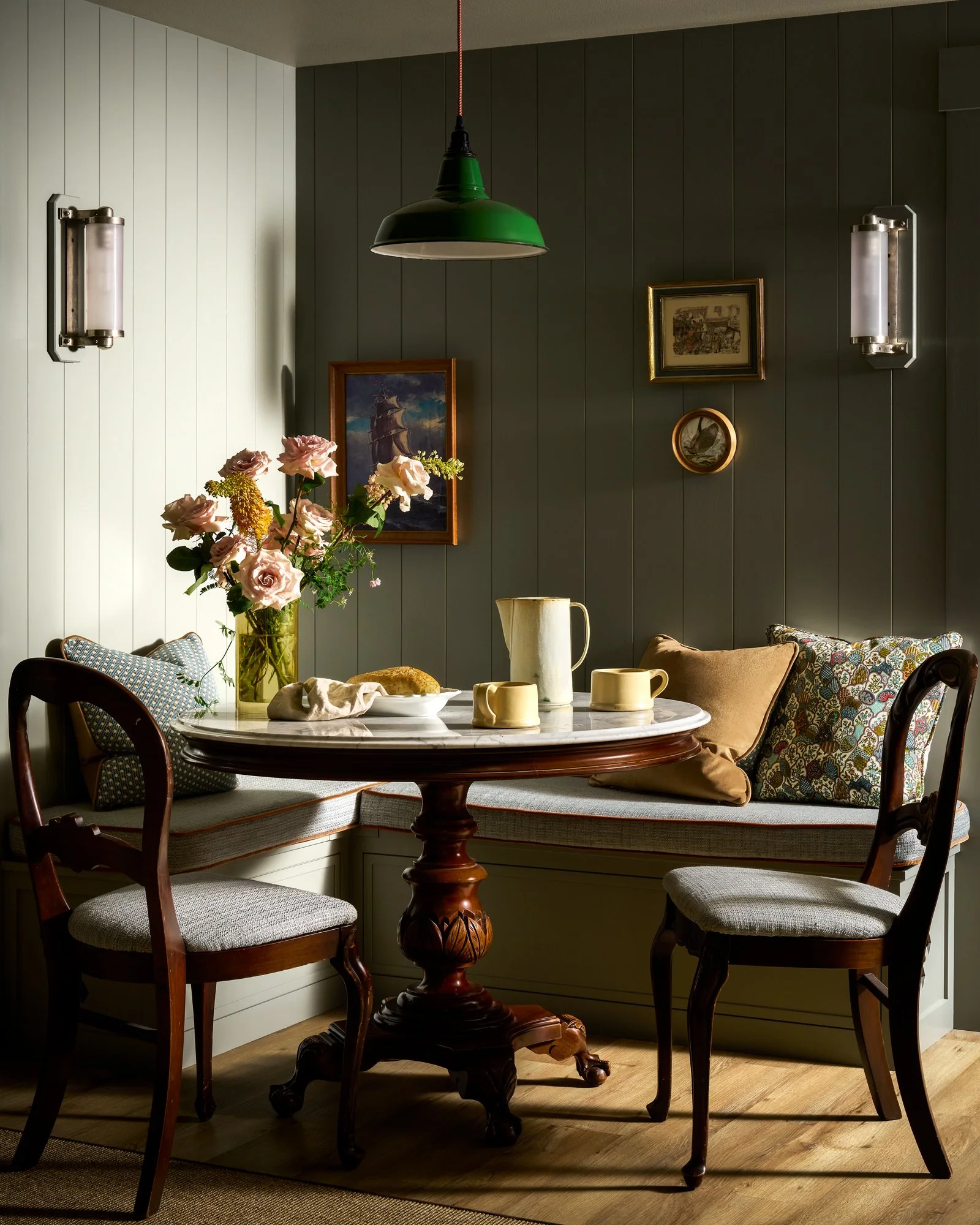 Elegant dining nook with a marble-top pedestal table, floral centerpiece, tea set, upholstered chairs, framed artwork, and green pendant light.