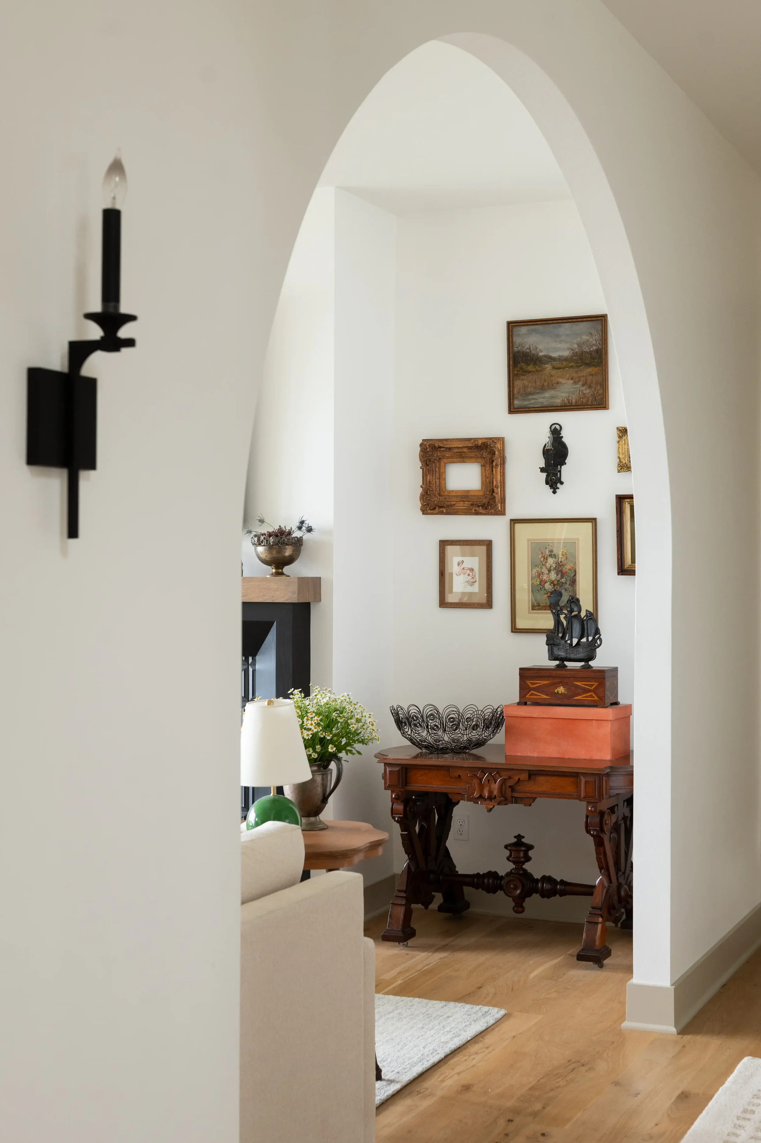 Elegant living room with arched doorway, antique artwork, ornate wooden console table, decorative objects, and a black wall sconce.