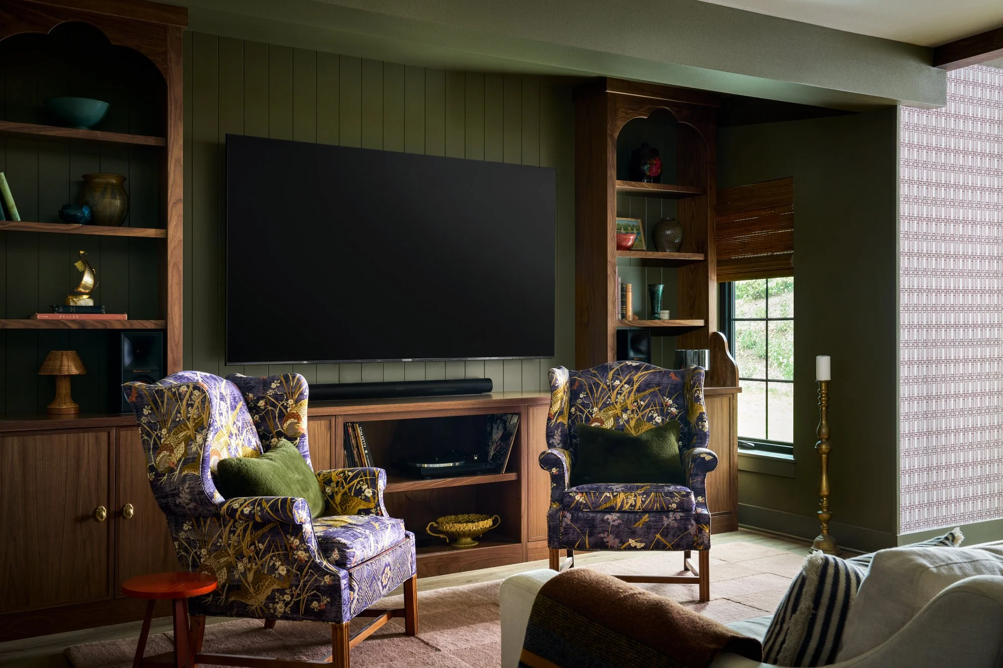 Living room with wooden shelves containing decorative vases and books, featuring a wall-mounted flat-screen TV, floral-patterned armchairs with green pillows, a window with brown blinds, and a candlestick next to a beige sofa.