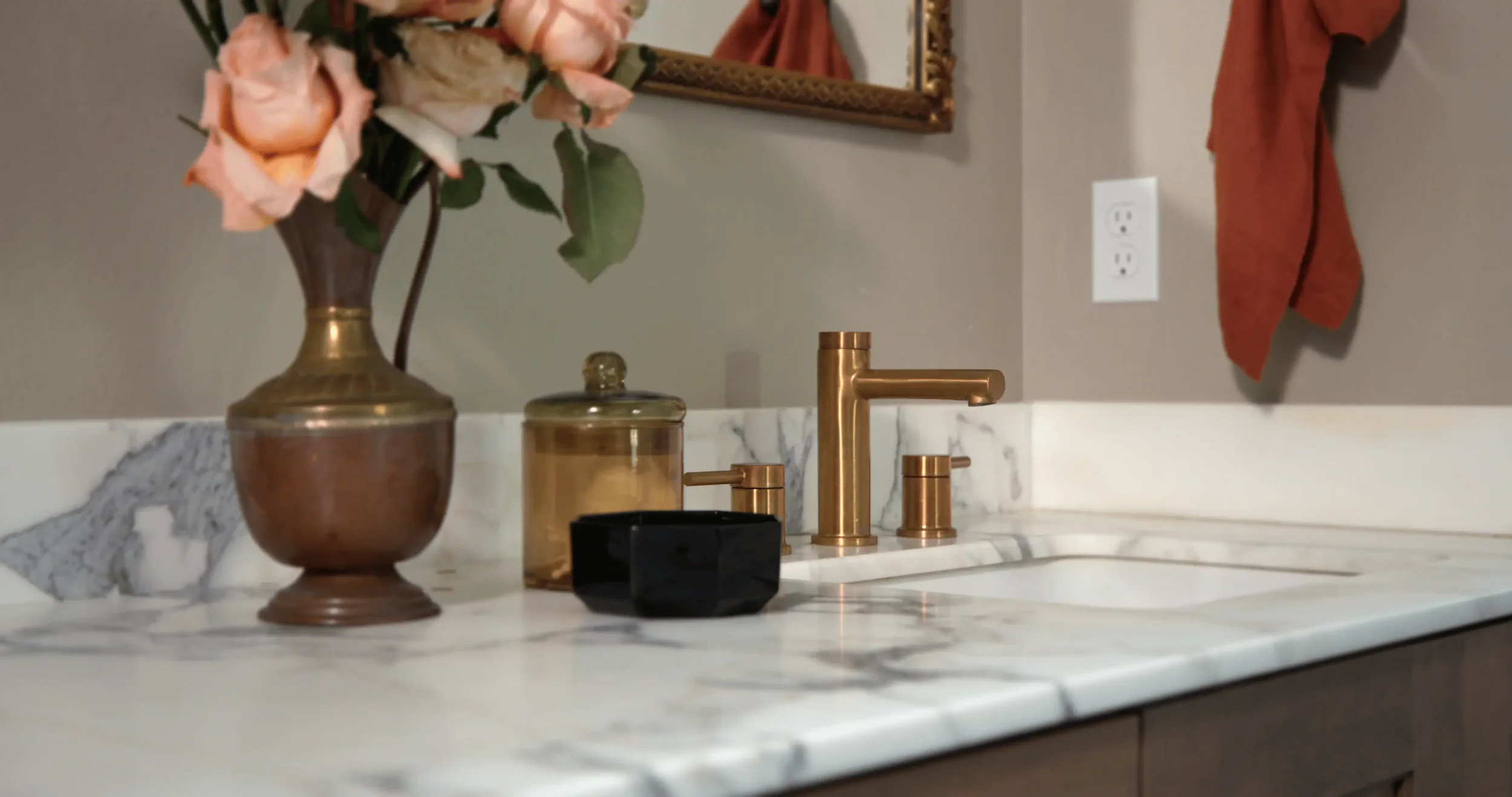 Marble bathroom vanity with brushed brass faucet, soap dispenser, copper vase of pink roses, and framed mirror