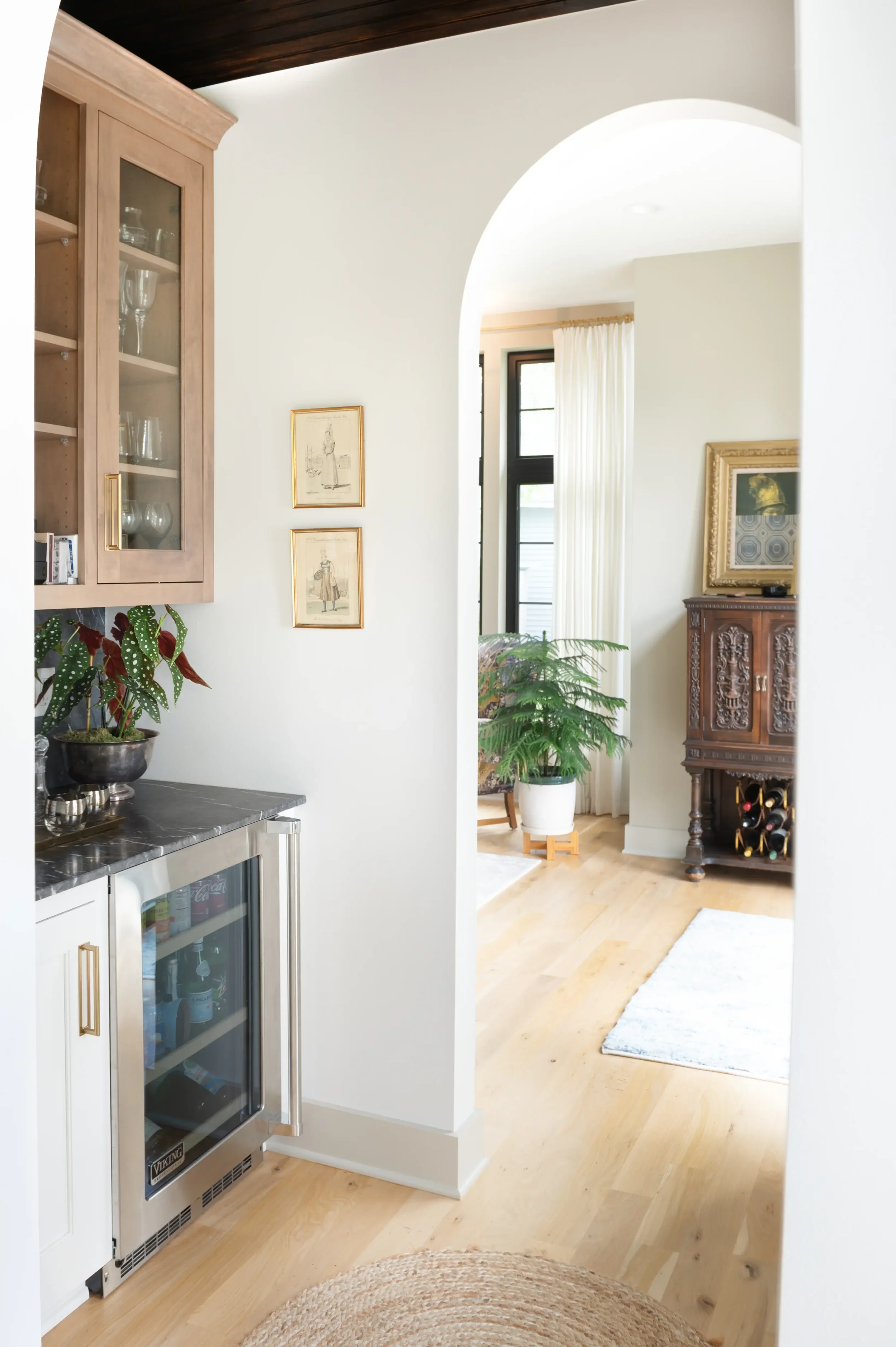 Bright home interior with a glass-front beverage refrigerator, wooden cabinetry, plants, framed artwork, and an arched doorway leading to a living room with a fireplace.