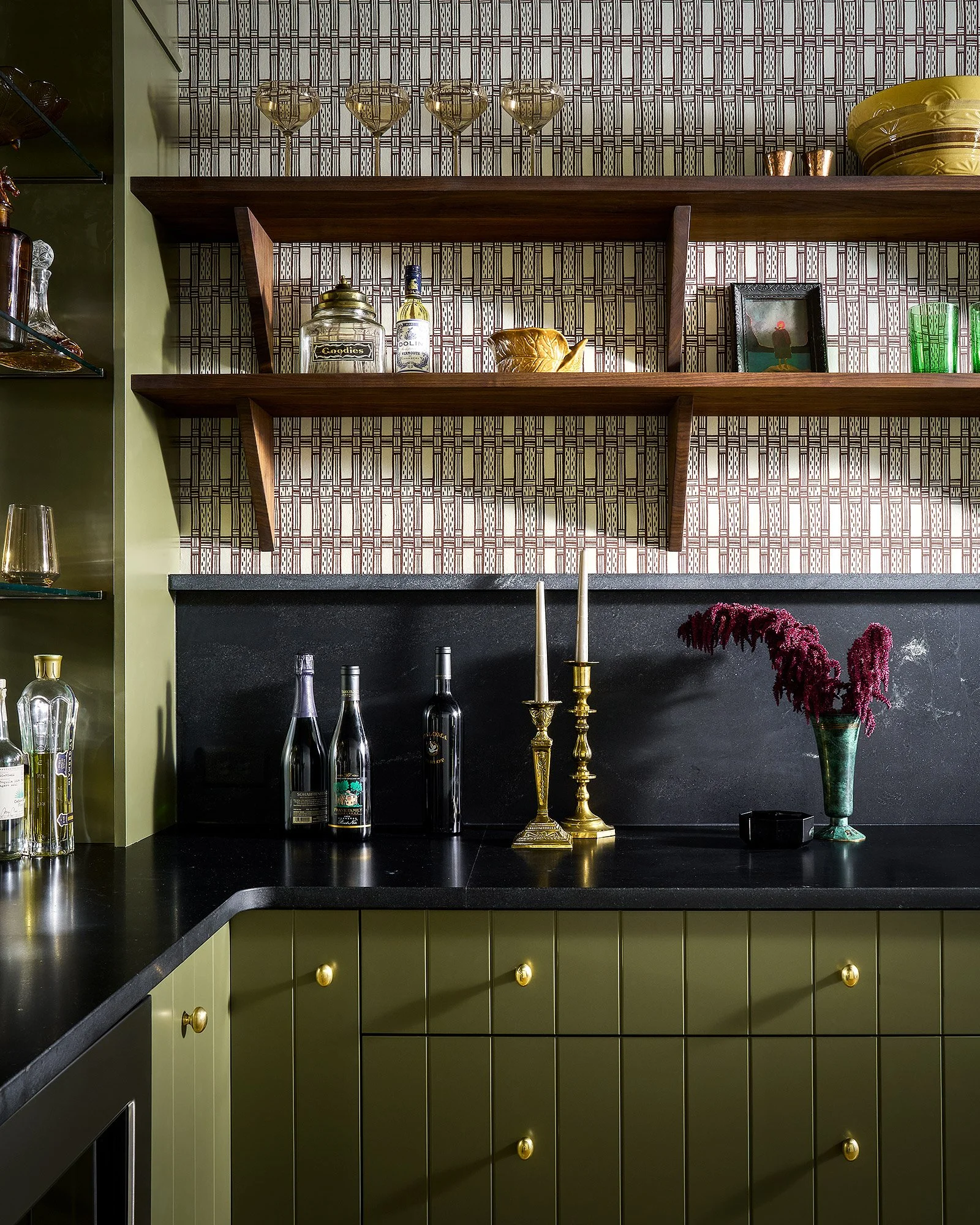 Green kitchen cabinets with black countertops, and open wooden shelves containing bottles, vases, and decorative items, set against patterned wallpaper.