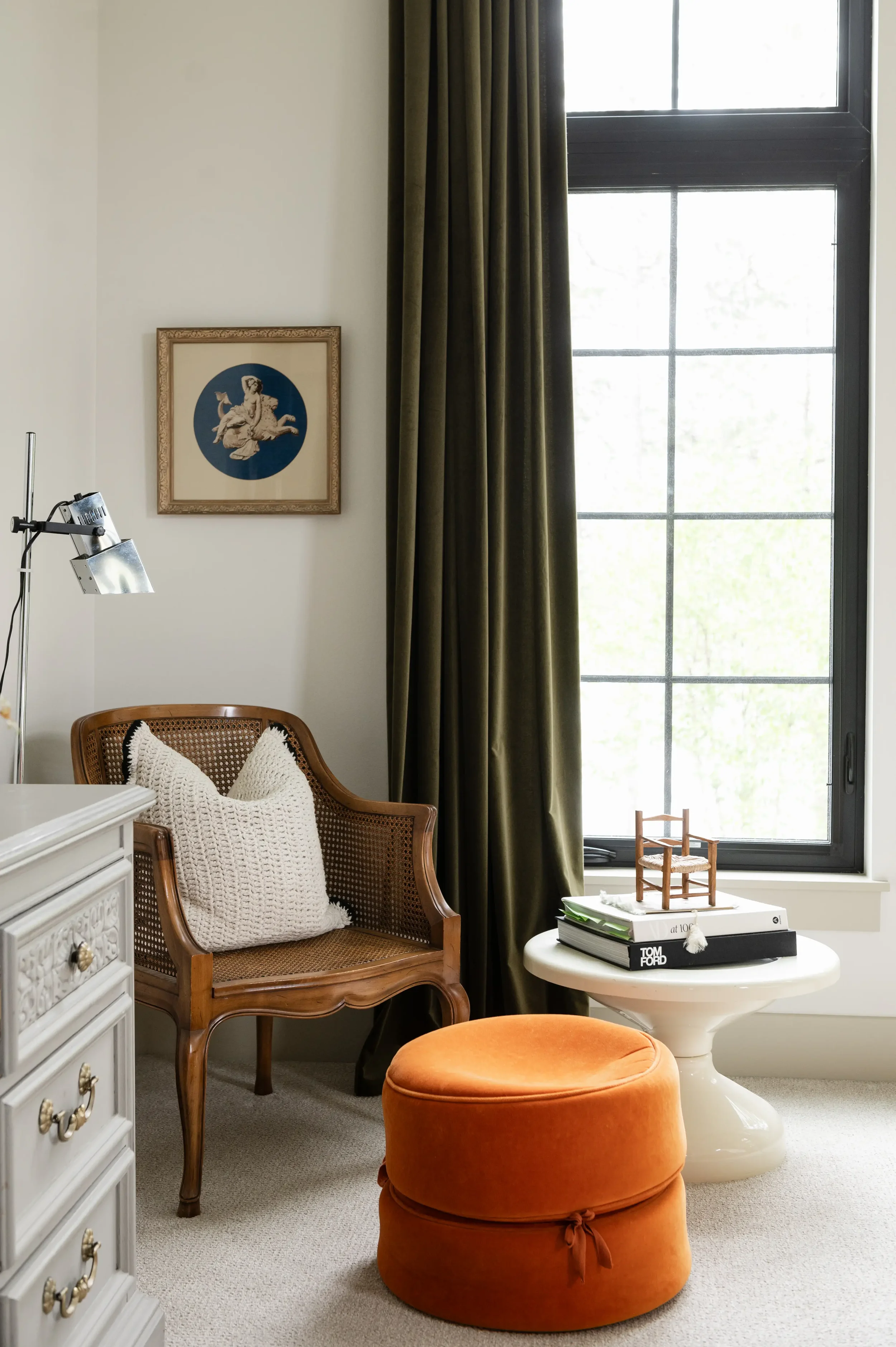 Bright reading nook with a cane armchair, white pillows, orange ottoman, floor lamp, framed artwork, dark curtains, and a round table beside a large window.