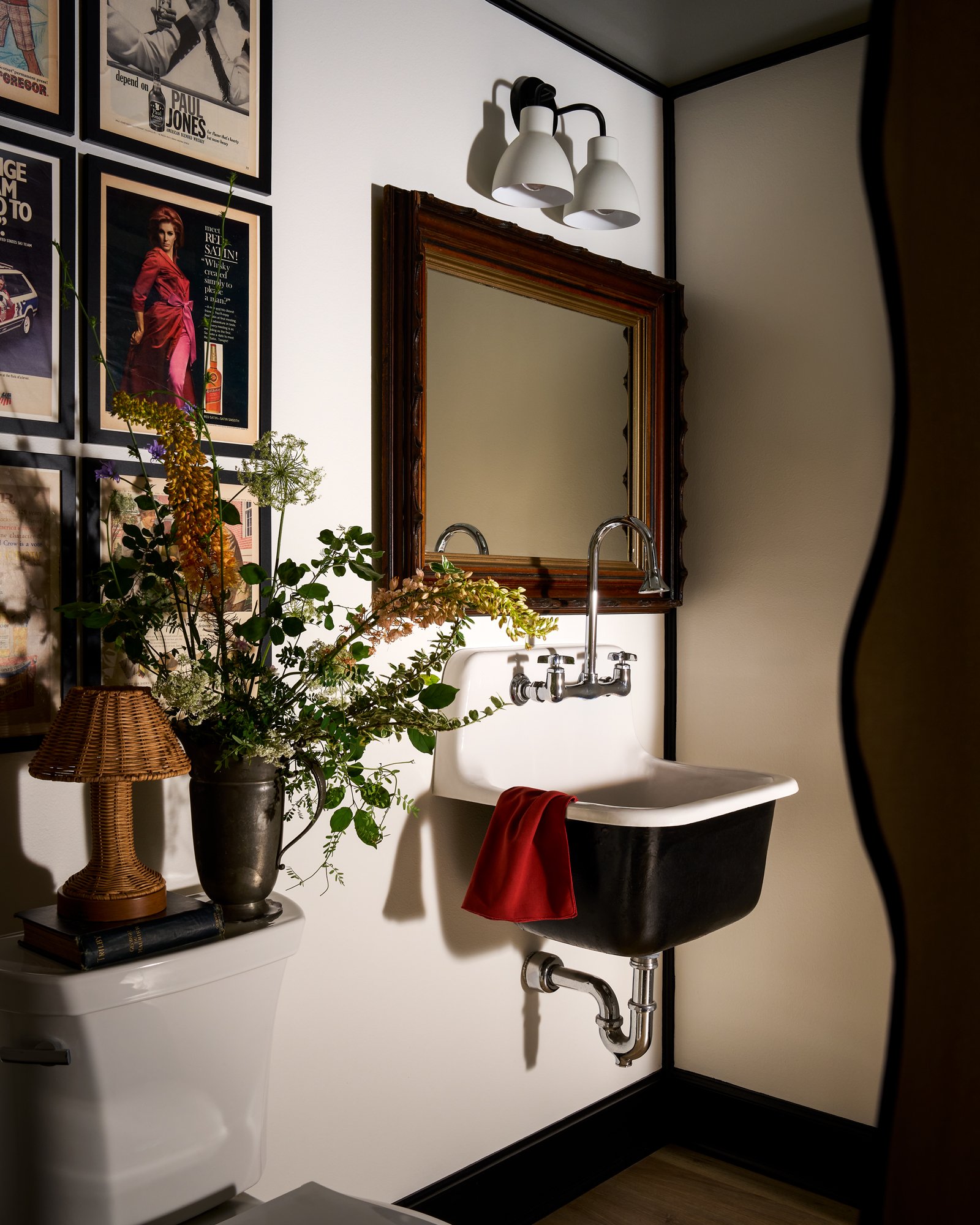 A bathroom decor scene with framed vintage posters on the wall, a large mirror with a wooden frame, a black and white vintage sink with a red cloth hanging from it, a small white side table with a woven lamp, and a vase with green and purple flowers.