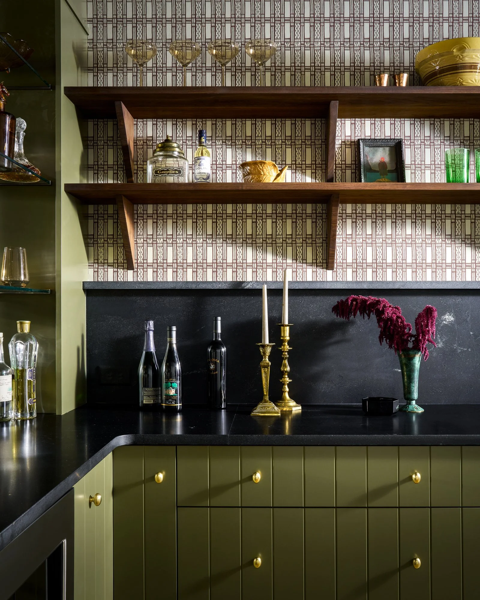 Stylish home bar with green cabinets, patterned wallpaper, open wood shelves, wine bottles, cocktail glasses, brass candlesticks, and decorative objects