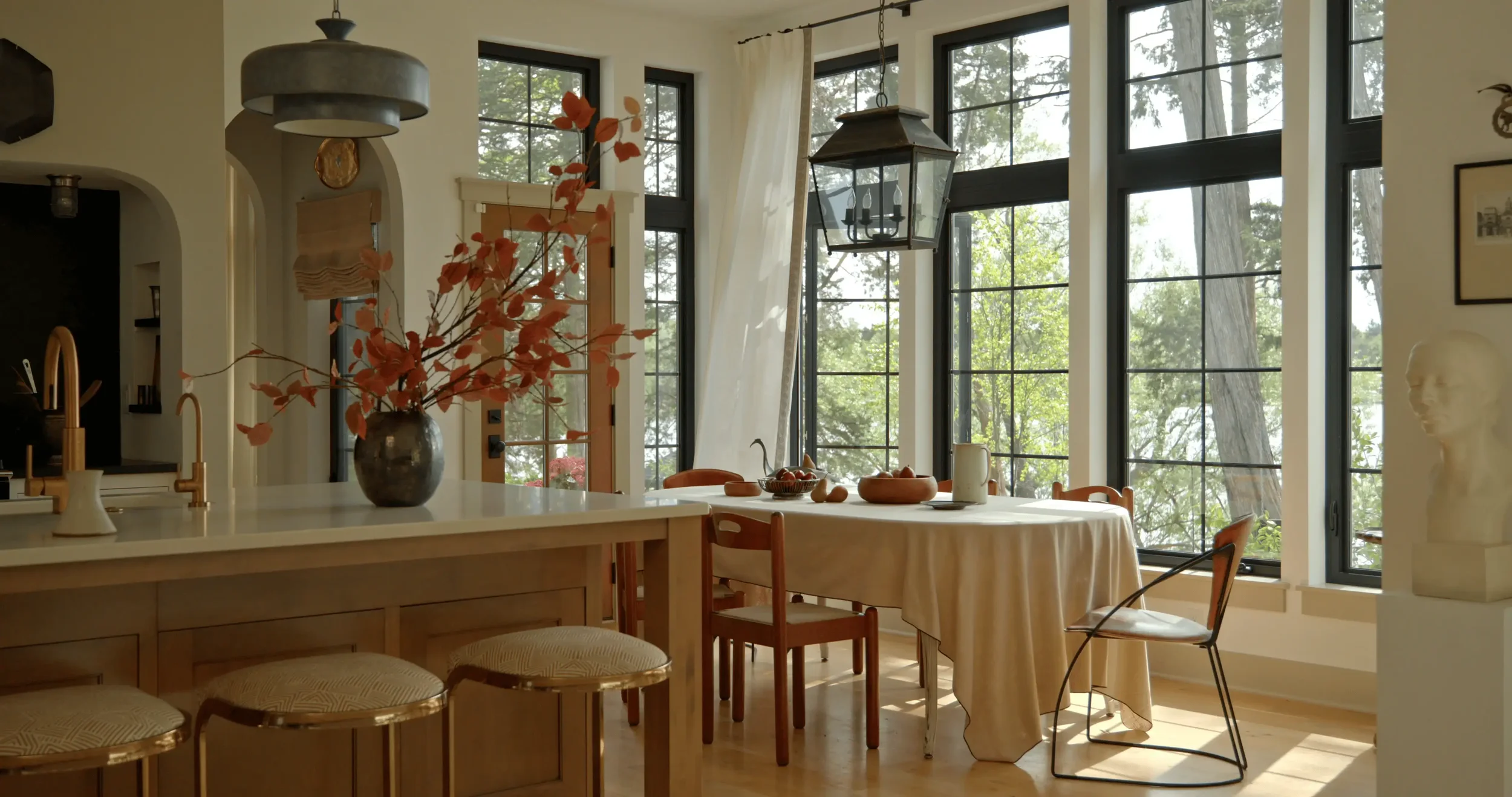 Sunlit open-plan kitchen and dining room with large black-framed windows, wood island, floral vase, round dining table, and hanging lantern