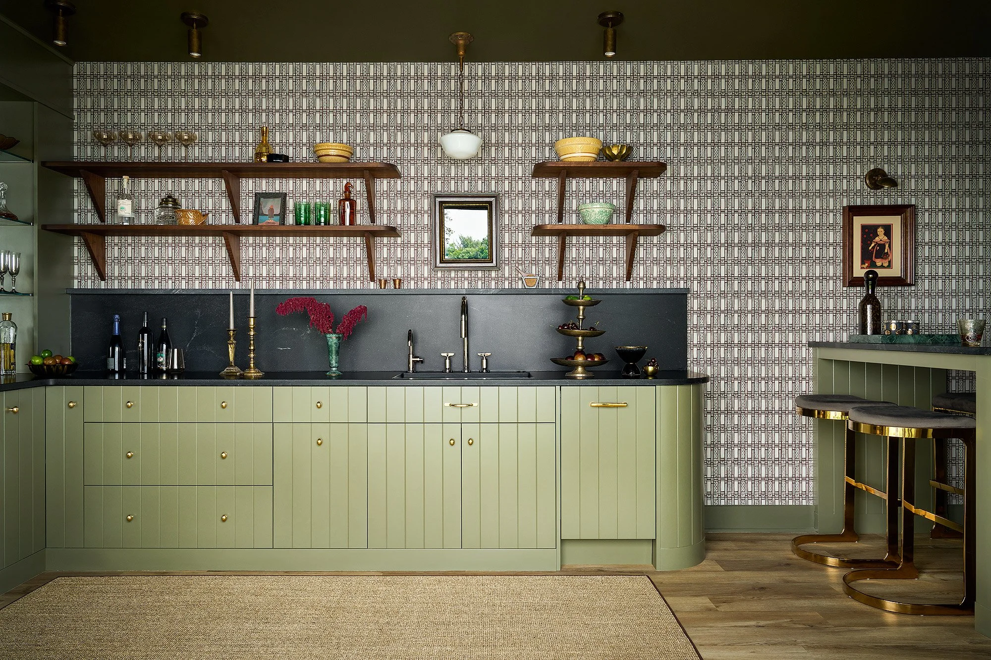 A modern kitchen with green cabinetry, black countertops, and open wooden shelves on a patterned wallpapered wall. Decorative items, glassware, and framed artwork are displayed, with a small window showing a view of trees outside.