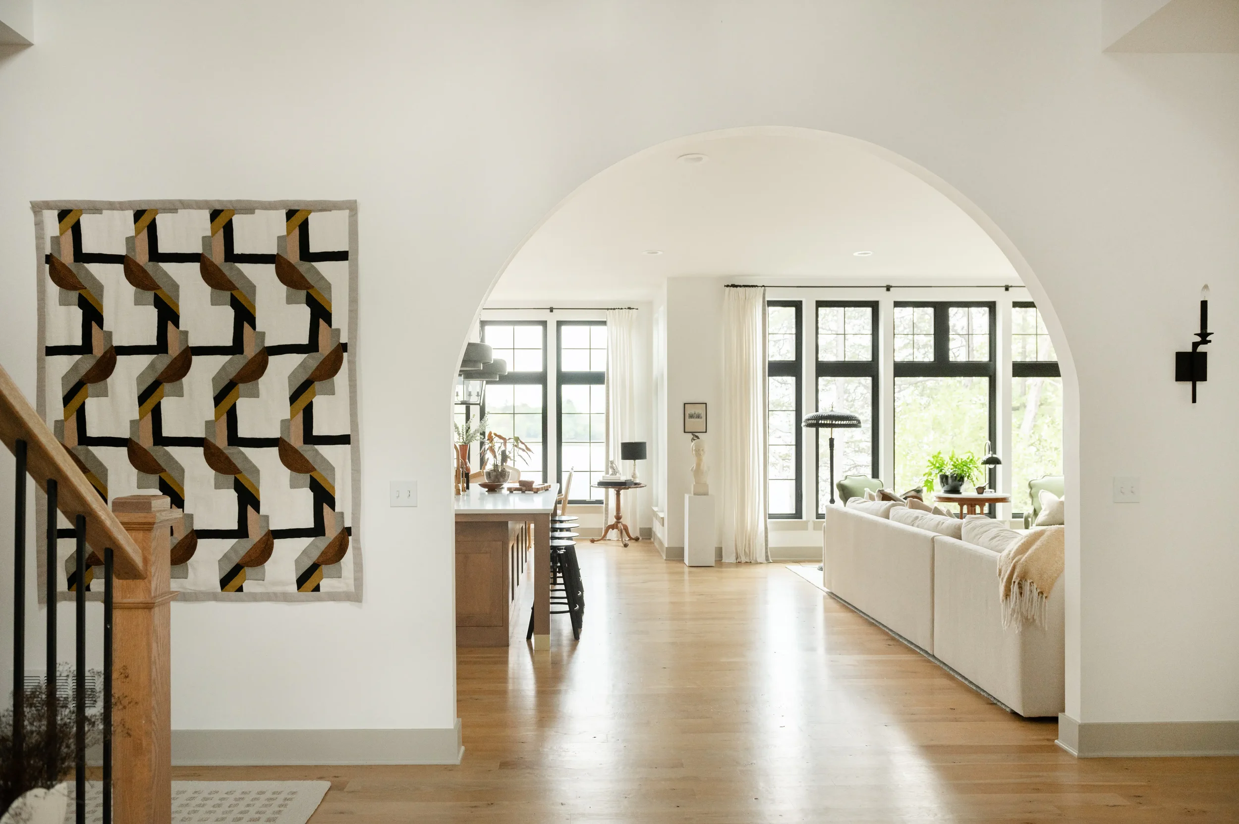 Bright modern open-plan home interior with arched doorway, geometric wall tapestry, staircase, hardwood floors, kitchen island, and large black-framed windows.