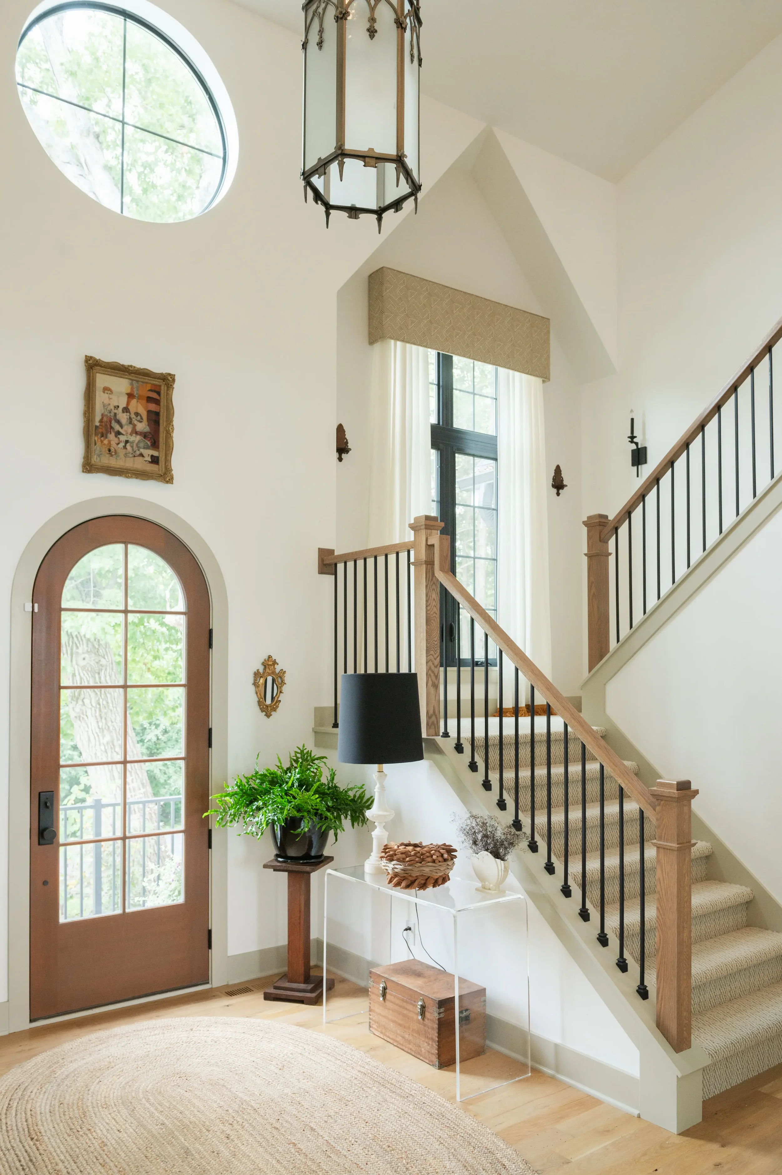 Bright modern entryway with a curved glass front door, staircase with black iron railings, large windows, pendant lantern, artwork, and indoor plants.