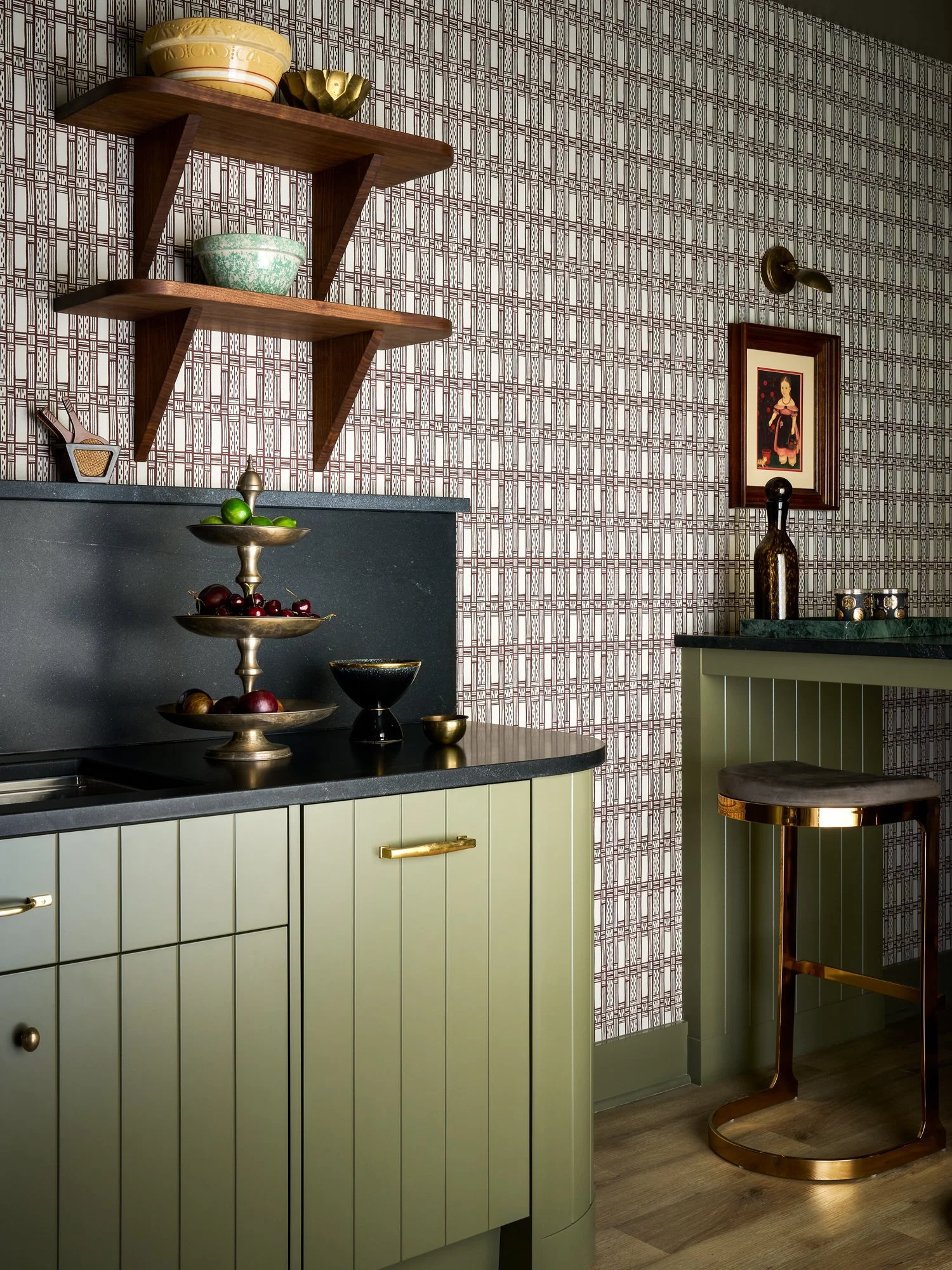 A kitchen with light green cabinets, a dark countertop, floating wooden shelves with bowls, a tiered fruit tray with cherries and limes, a framed artwork on the wall, and a modern bar stool with a gold base.