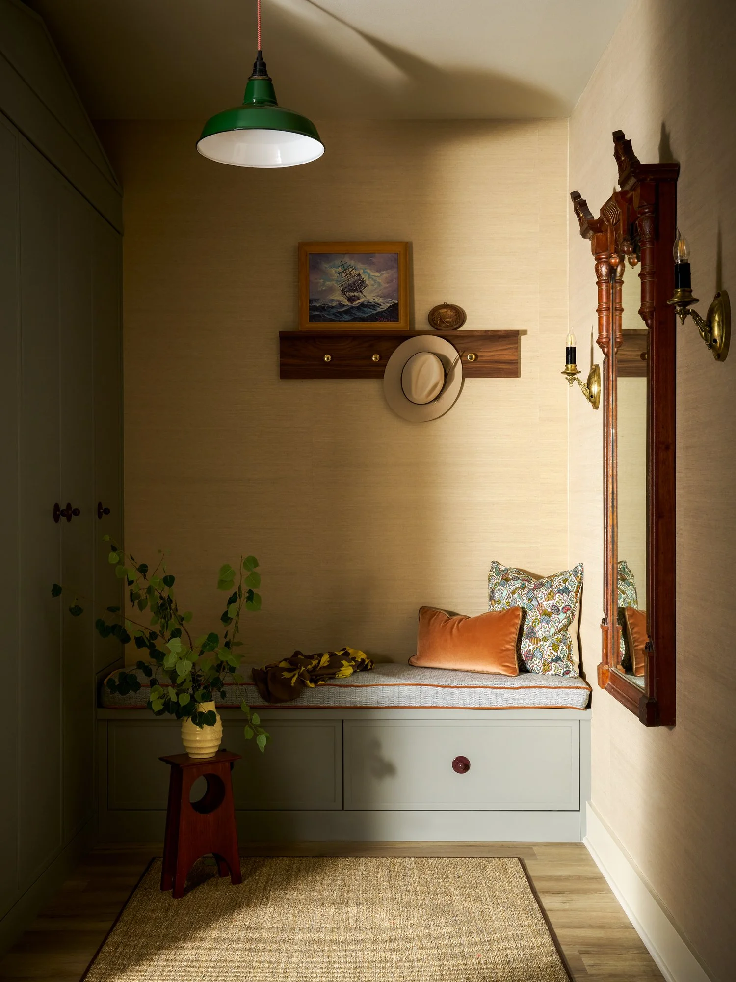 A cozy corner with a built-in bench seat topped with colorful pillows, a small wooden table with a potted plant, a large mirror, a wooden shelf holding a framed painting of a ship caught in a storm, and a wall-mounted hat and coat rack. The room has beige walls, wooden flooring, and a green ceiling lamp.