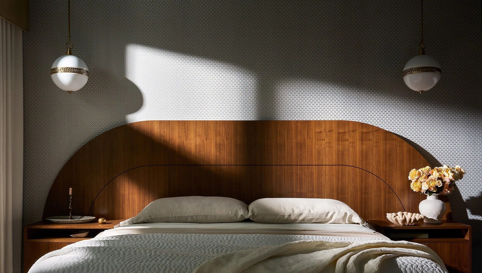 A bedroom with a wooden headboard, wall-mounted lamps, and a textured wallpaper. There is a bedside table on each side of the bed, one with a candle and decorative object, and the other with a white vase holding pink and white flowers. Sunlight creates a bright patch on the wallpaper.