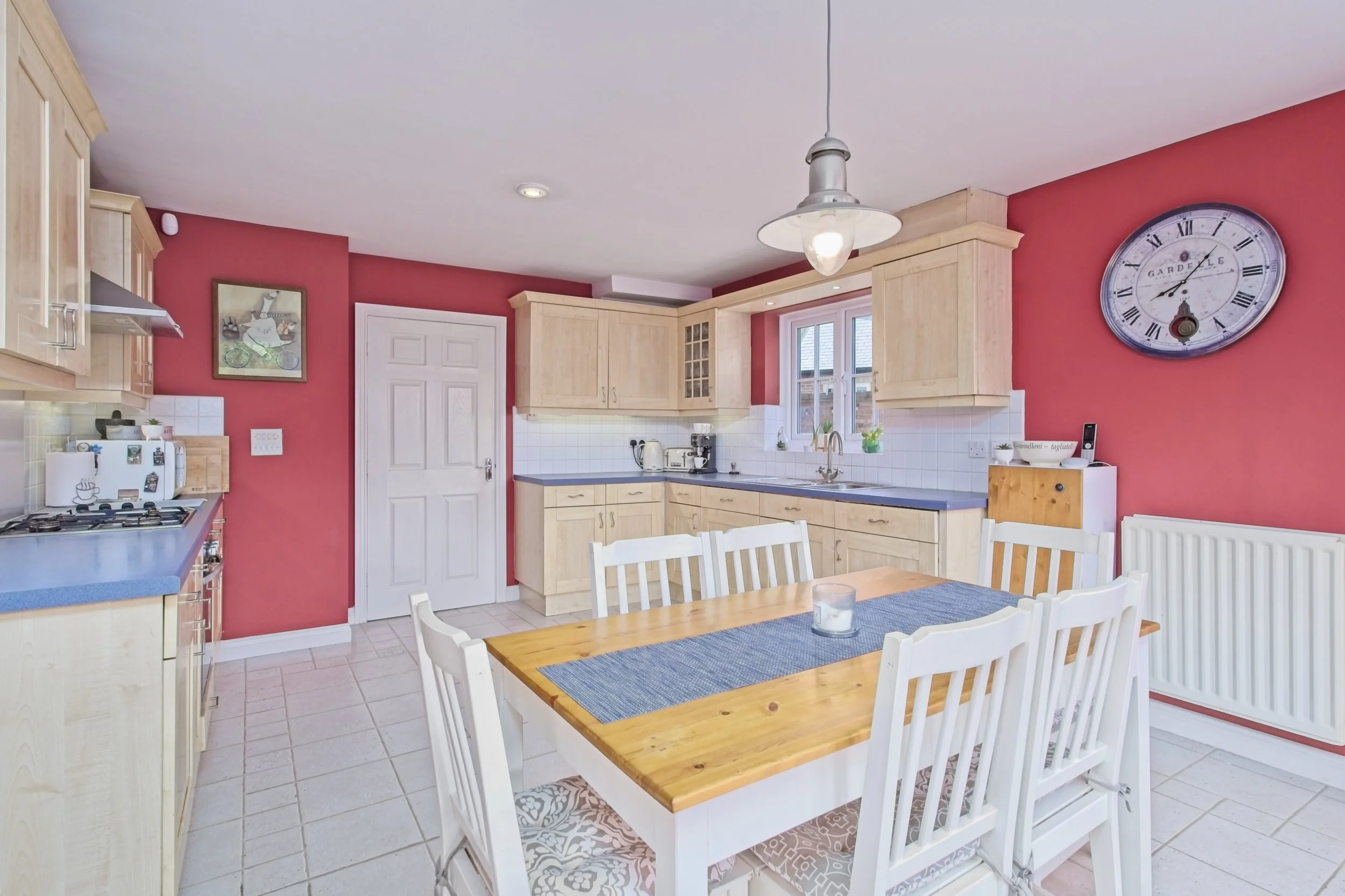 Kitchen with red walls, light wood cabinets, white chairs around a wooden table, and a large wall clock.
