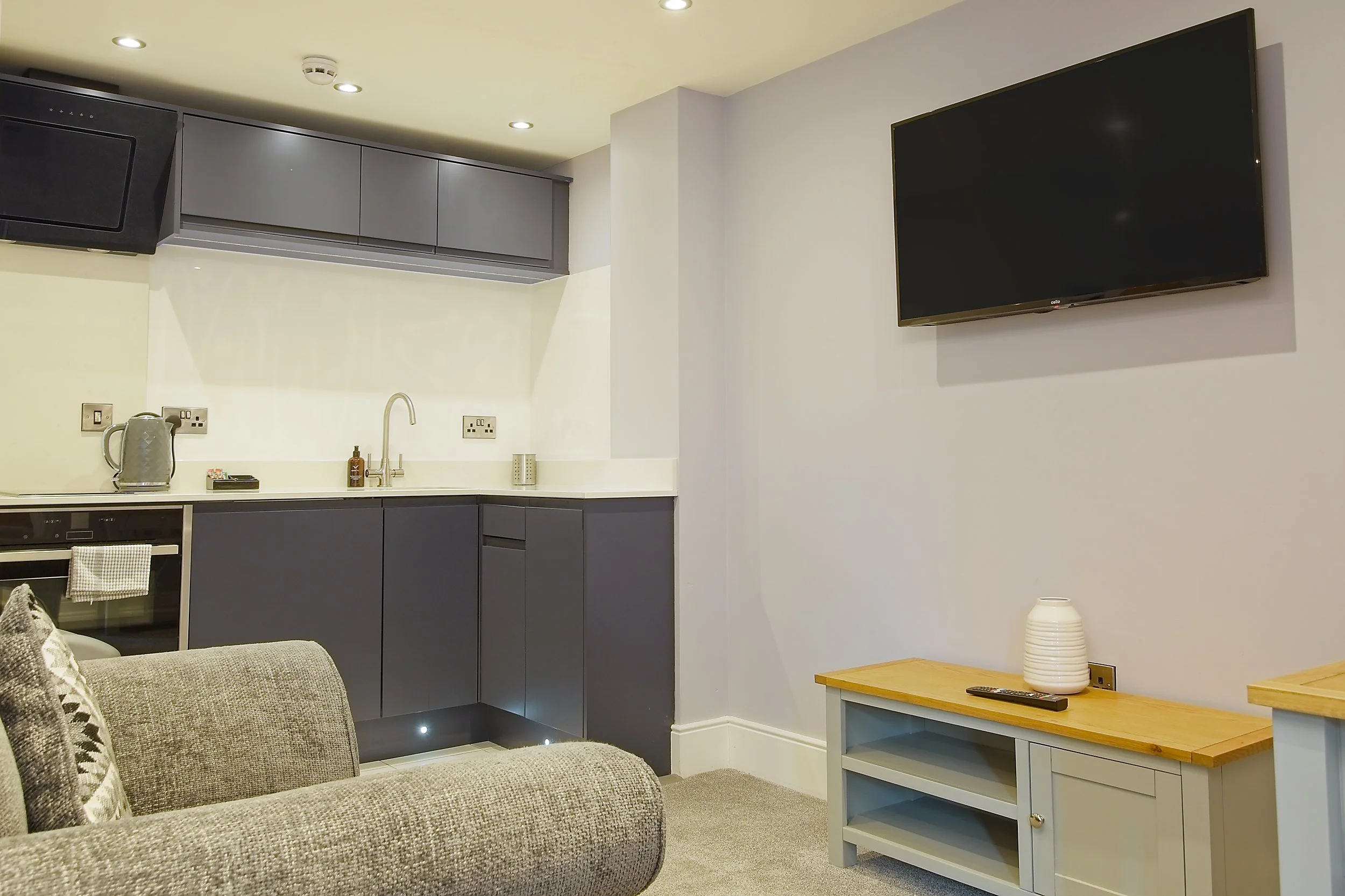 A living room with a kitchen area. The kitchen has dark gray cabinets, a countertop with a kettle, soap dispenser, and a small tray. There is a sink with a faucet, and various electrical outlets on the wall. On the right side, a flat-screen TV is mou