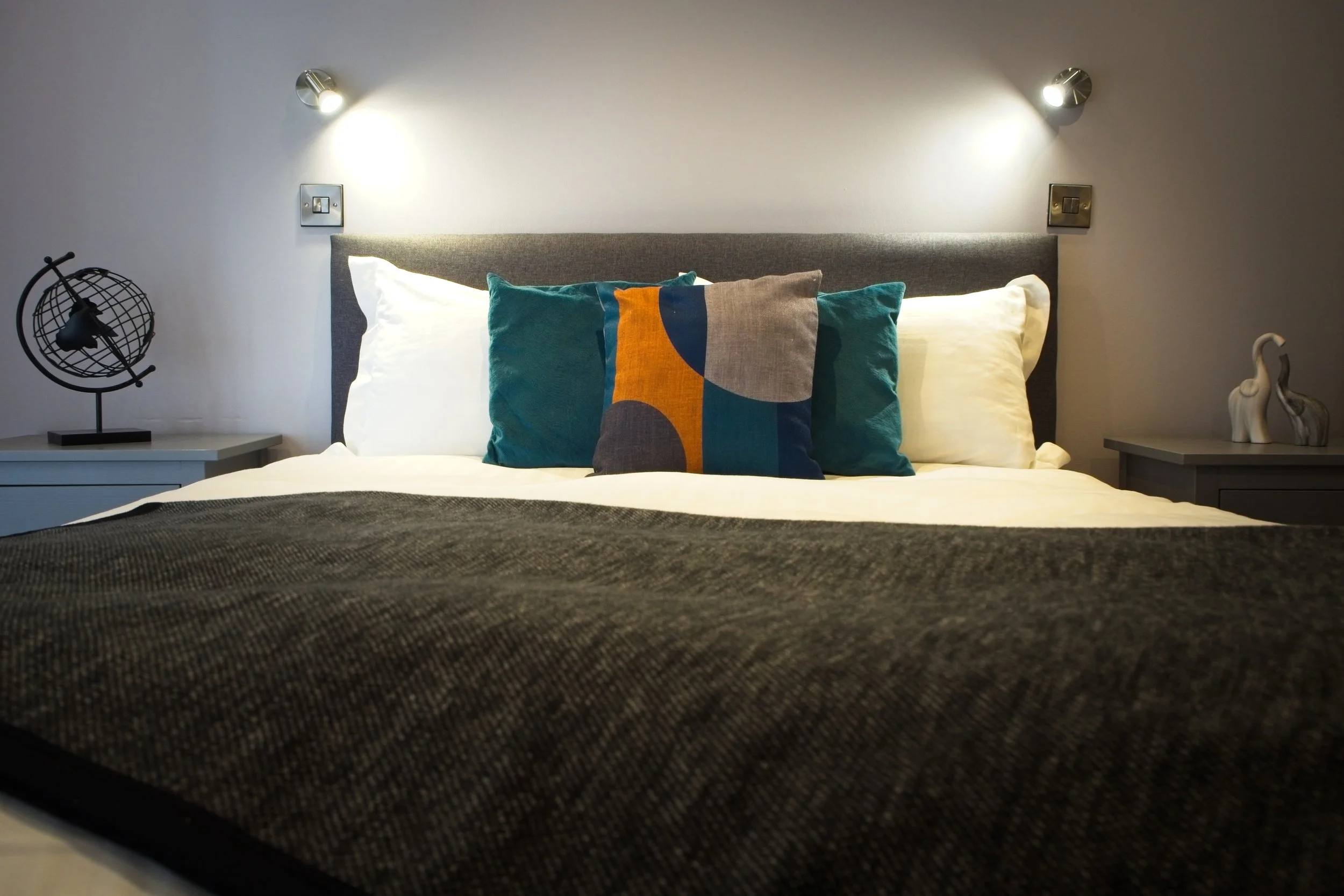 Close-up view of a neatly made bed with a dark textured blanket, decorative pillows with vibrant patterns, and two wall-mounted reading lights above.