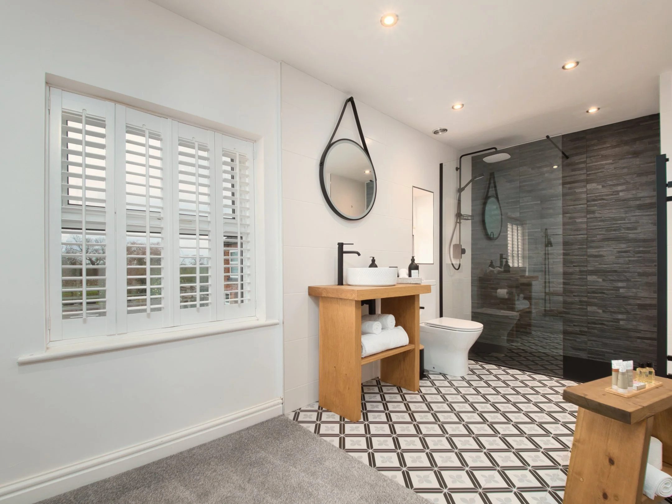 Modern bathroom with black and white patterned floor tiles, white walls, and a large window with white blinds. There is a vanity with a vessel sink, black fixtures, and a wooden shelf underneath holding towels. A mirror hangs on the wall, and a showe