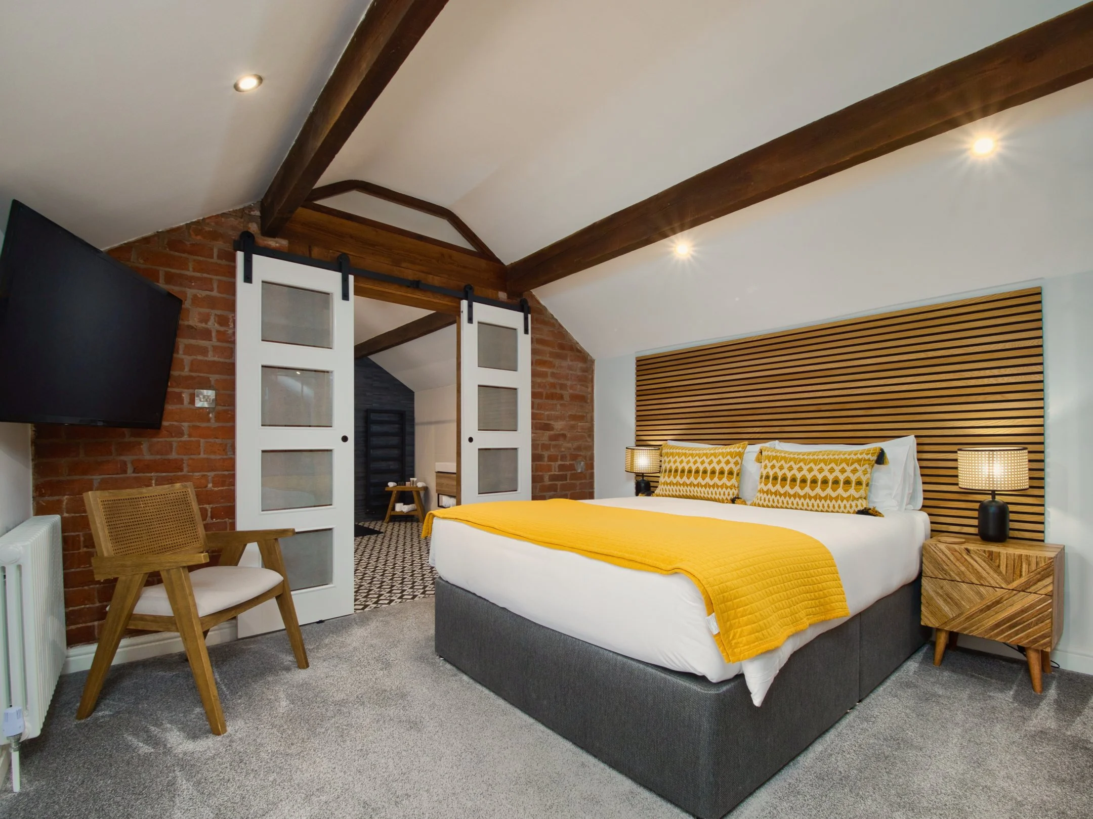 A modern bedroom featuring a bed with white bedding and a yellow quilt, two matching yellow and white patterned pillows, bedside tables with lamps, wooden slatted headboard, sliding barn-style doors, exposed wooden beams on the ceiling, a wall-mounted flat-screen TV, and brick accents.