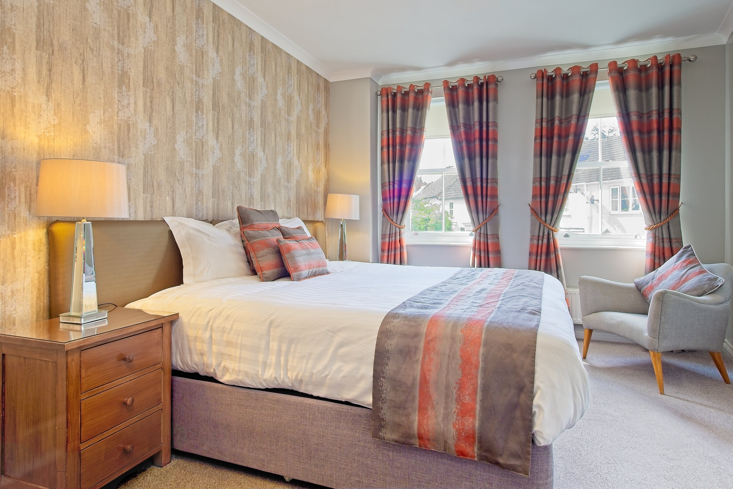 A cozy bedroom with a large bed, wooden nightstand, and a beige upholstered headboard. The bed has white linens and matching pillows with red and gray patterns. Two bedside lamps with white shades illuminate the room. The wall behind the bed is cover