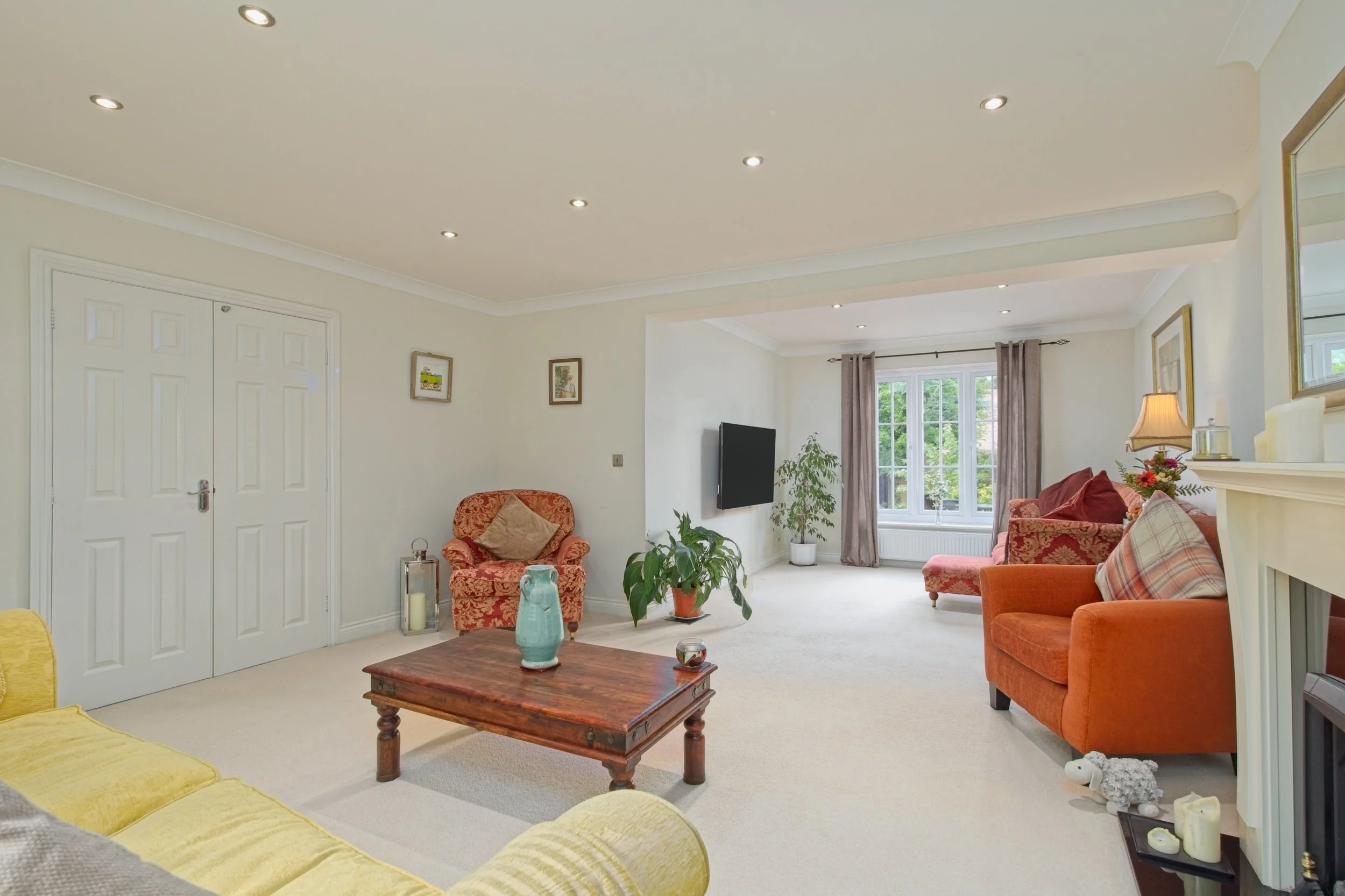 A spacious living room with cream-colored walls and ceiling, decorated with small framed artwork. There are two white double doors on the left, a flat-screen TV on the right wall, and a large window with beige curtains letting in natural light. The r