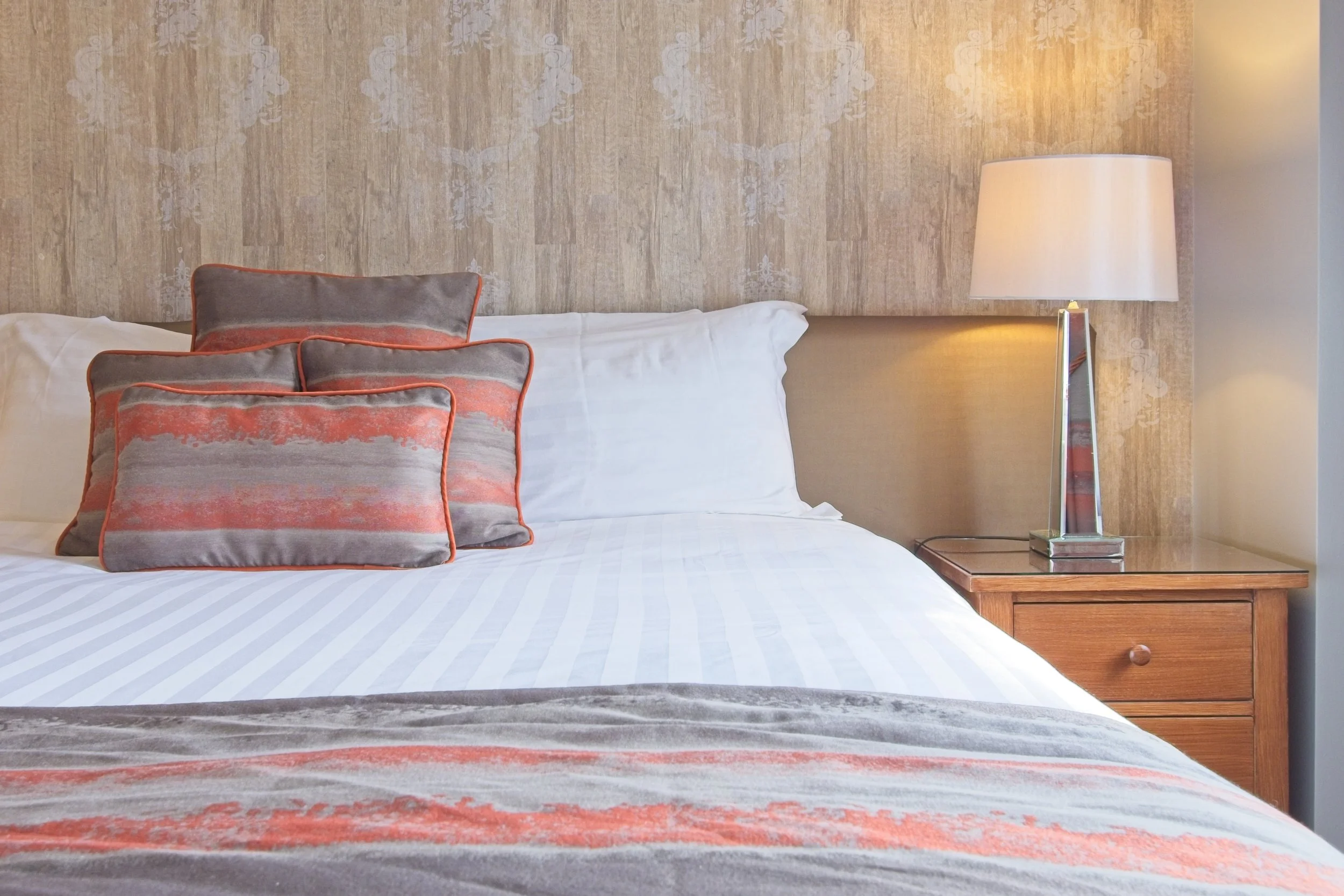 A neatly made bed with white bed sheets, two large white pillows, and a decorative pillow with a gray and coral pattern, in a hotel room with beige wallpaper and a wooden nightstand with a modern white lamp.