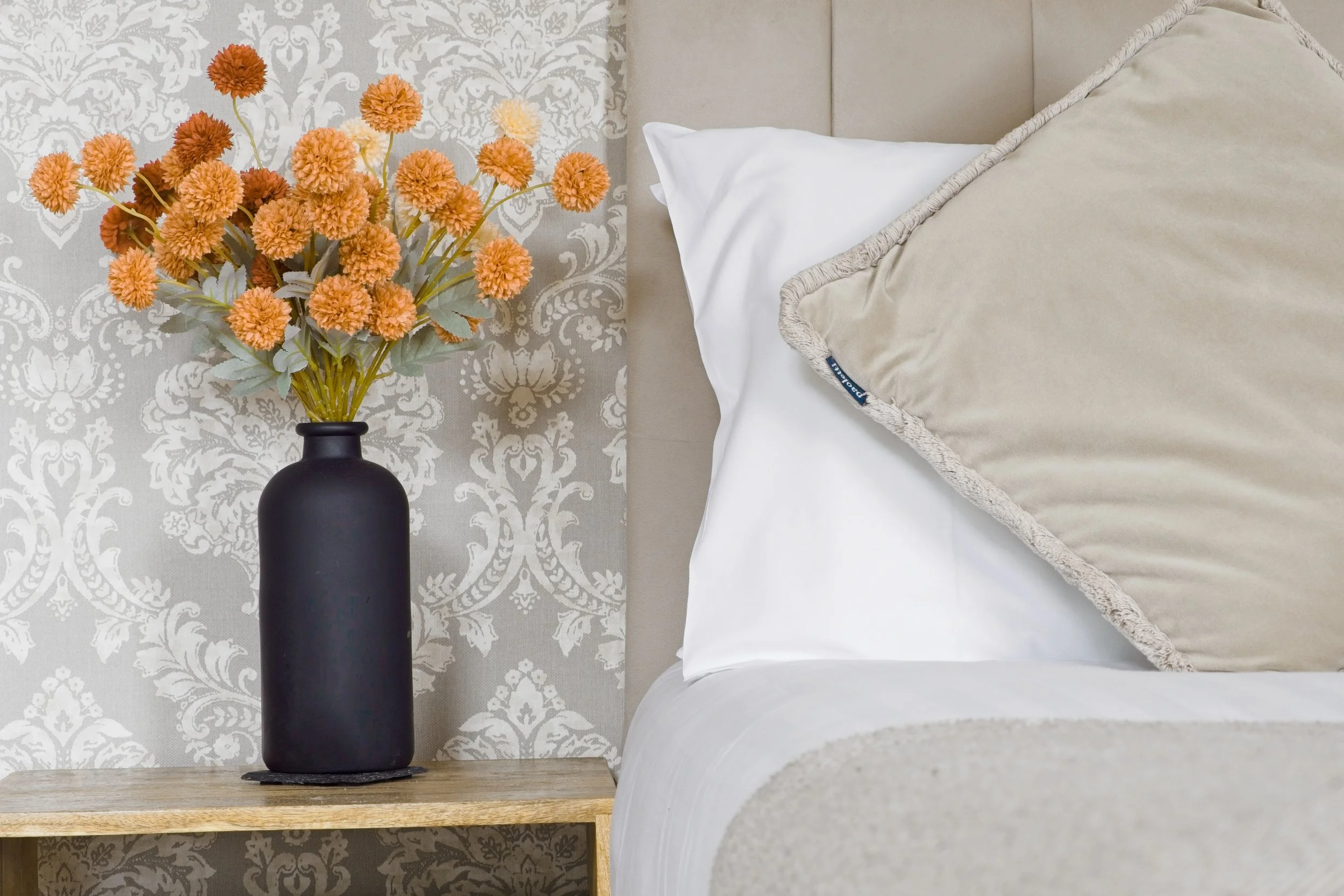 An example of a feature photo of a black vase with orange and yellow artificial flowers on a wooden nightstand beside a bed with white pillow and beige bedding, against a patterned wallpaper background.